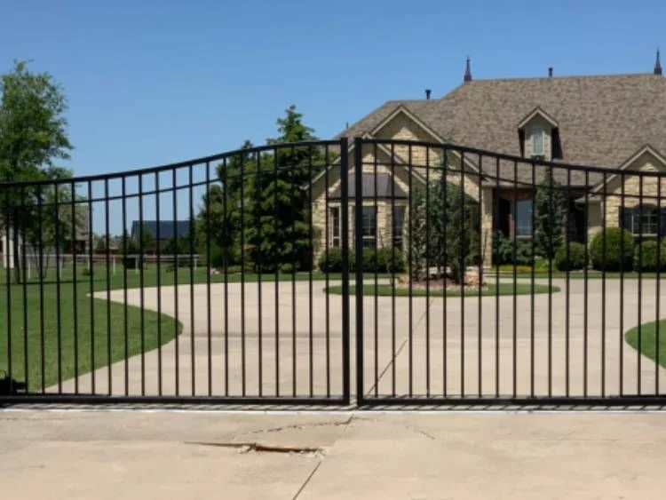 Arched black ornamental steel double driveway security gate installed at the concrete entrance of an executive stone home.