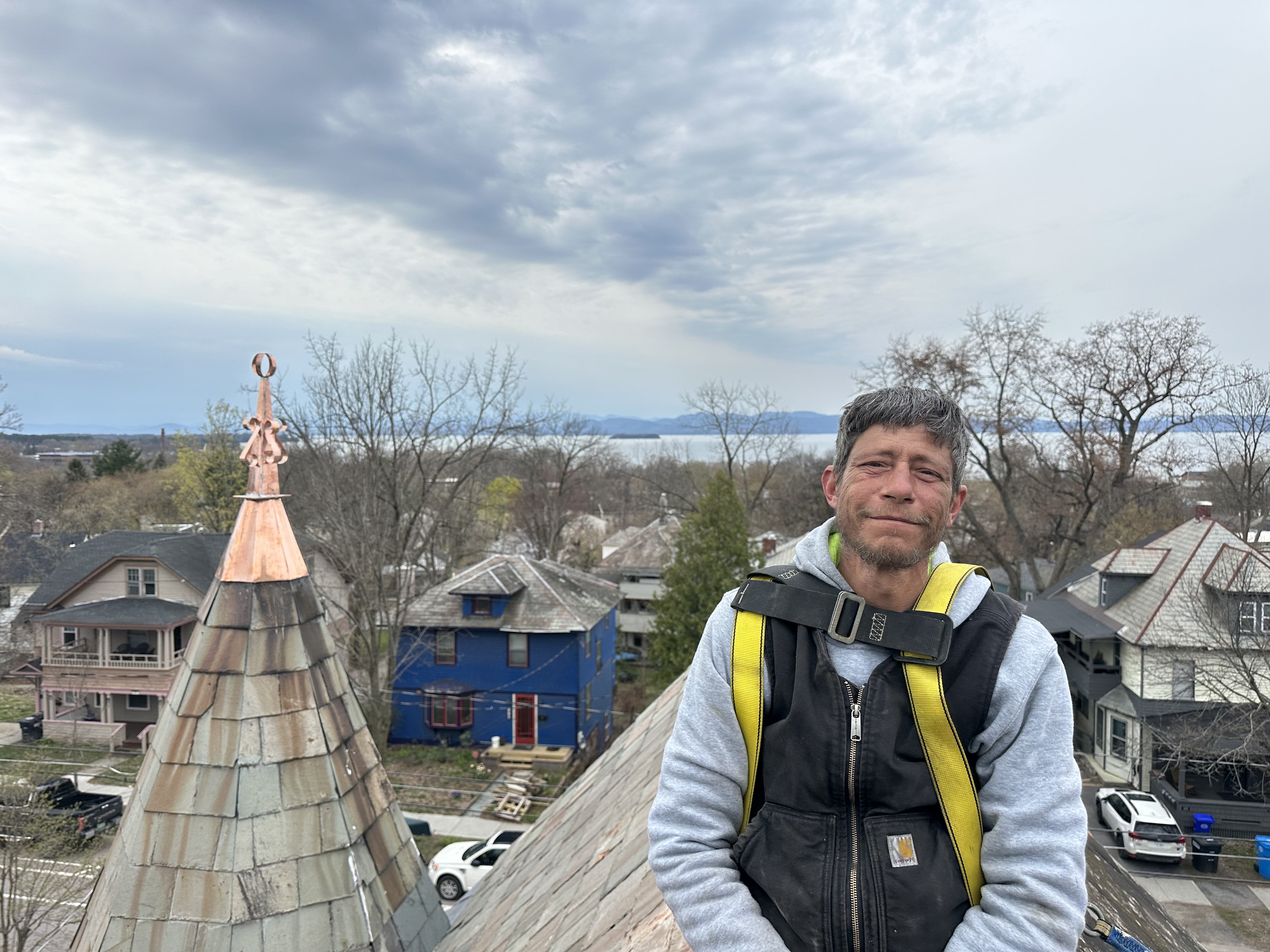 Roofer in safety harness atop slate roof with lake and mountain views