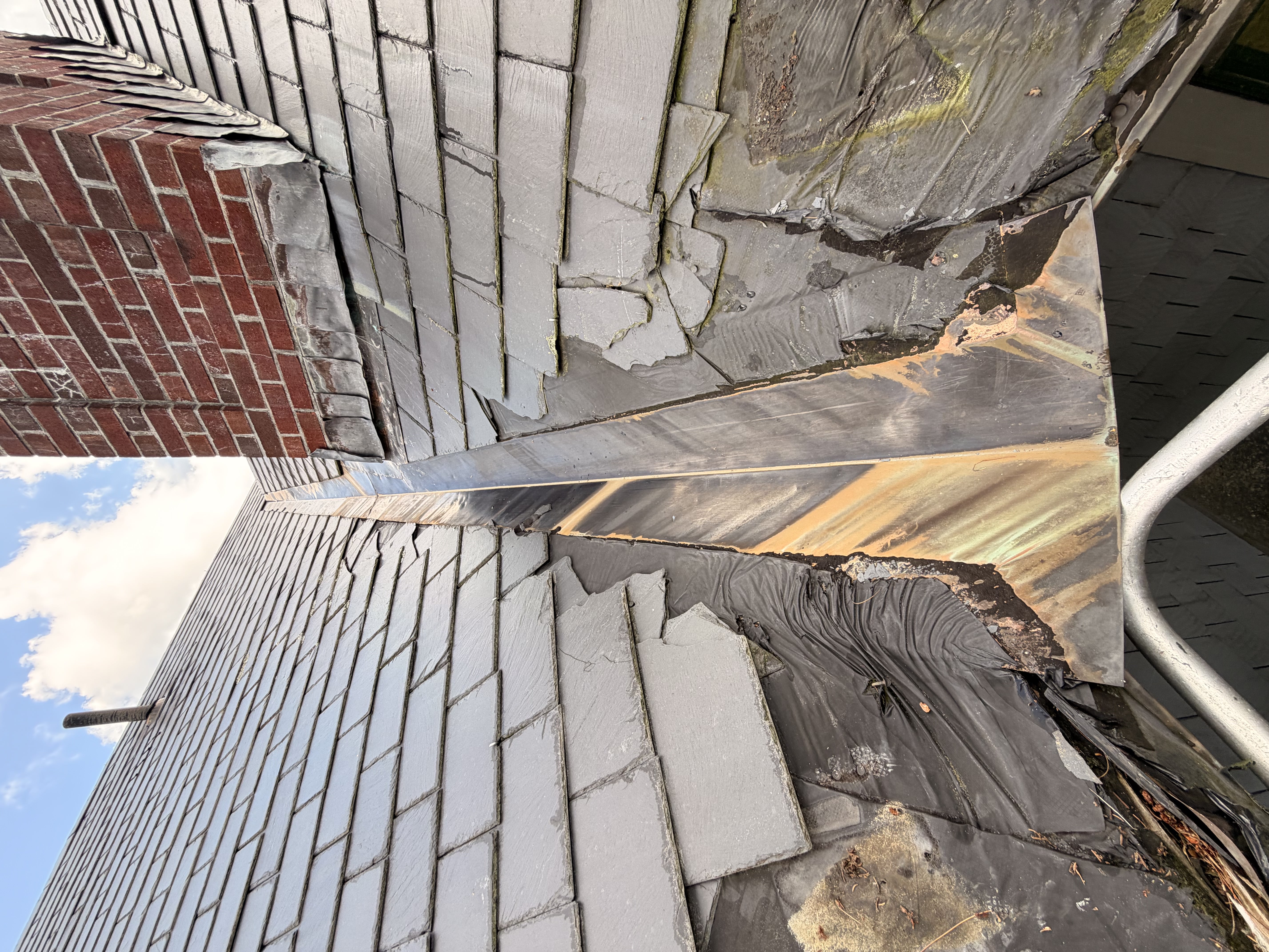 Failing roof edge with deteriorated slate, damaged flashing, and water damage on historic building