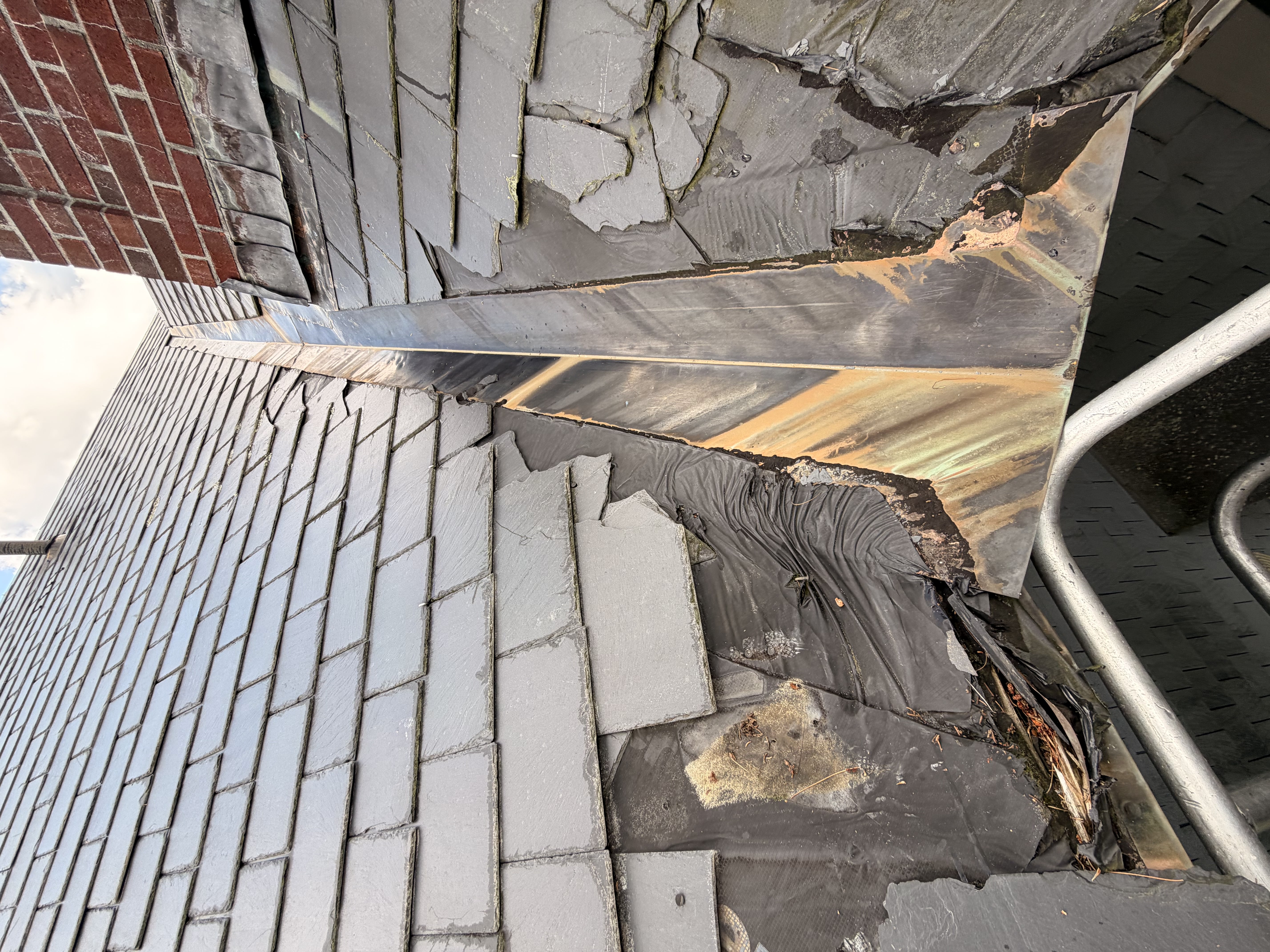 Deteriorated copper flashing and damaged slate tiles on historic home roof edge