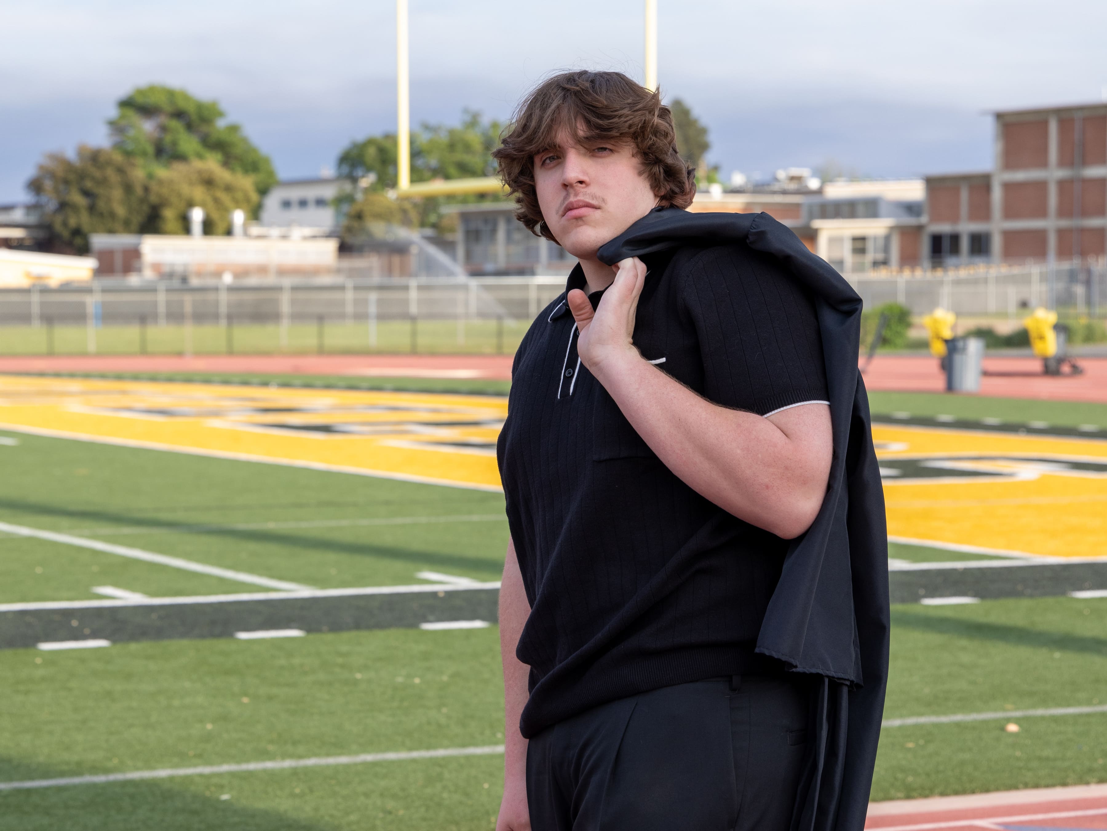 Senior in black polo with jacket over shoulder on football field by Brooke Lazzarini