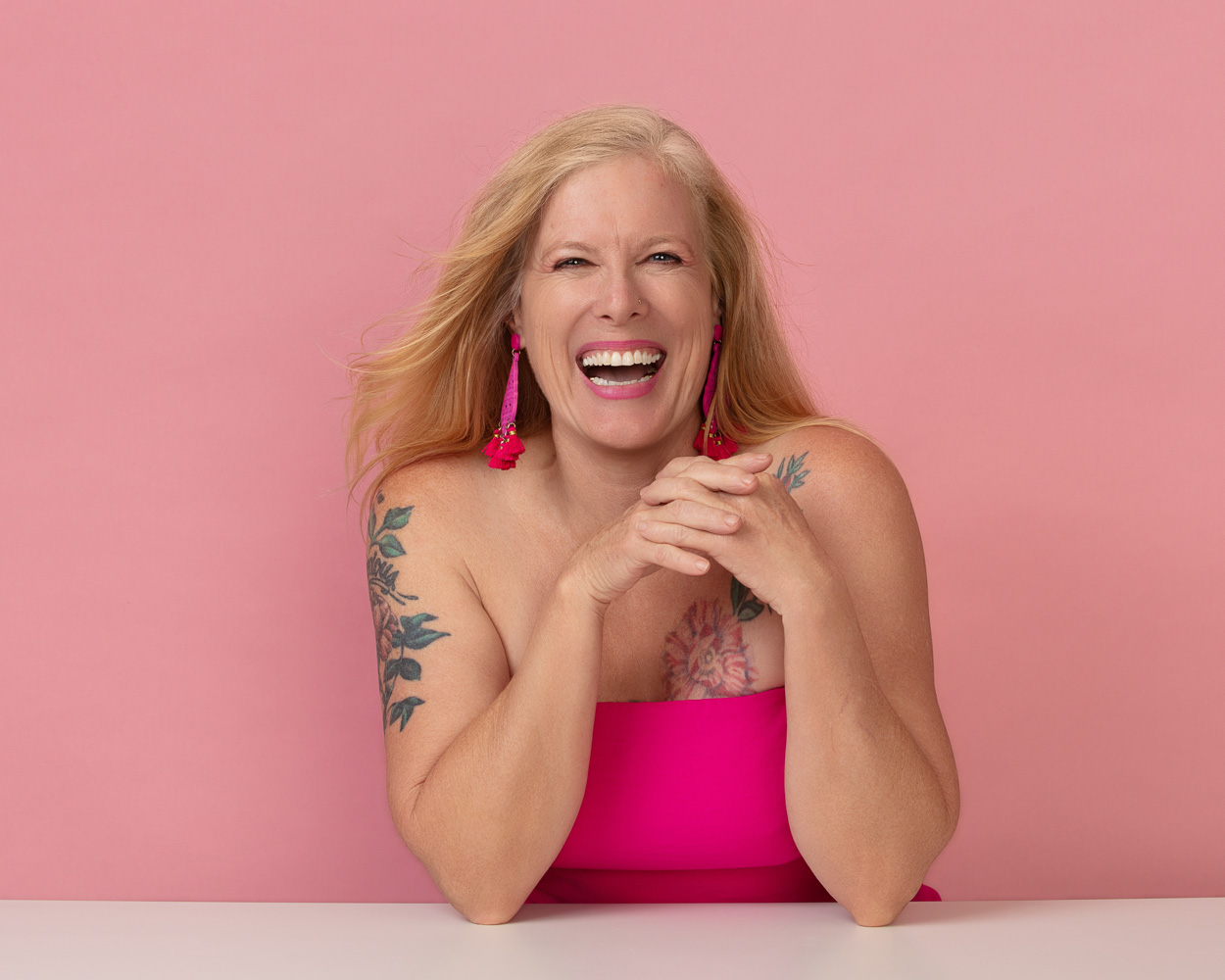 Morgan Daily Jackson in a bright pink strapless top against a pink background, with floral tattoos visible and hands clasped warmly
