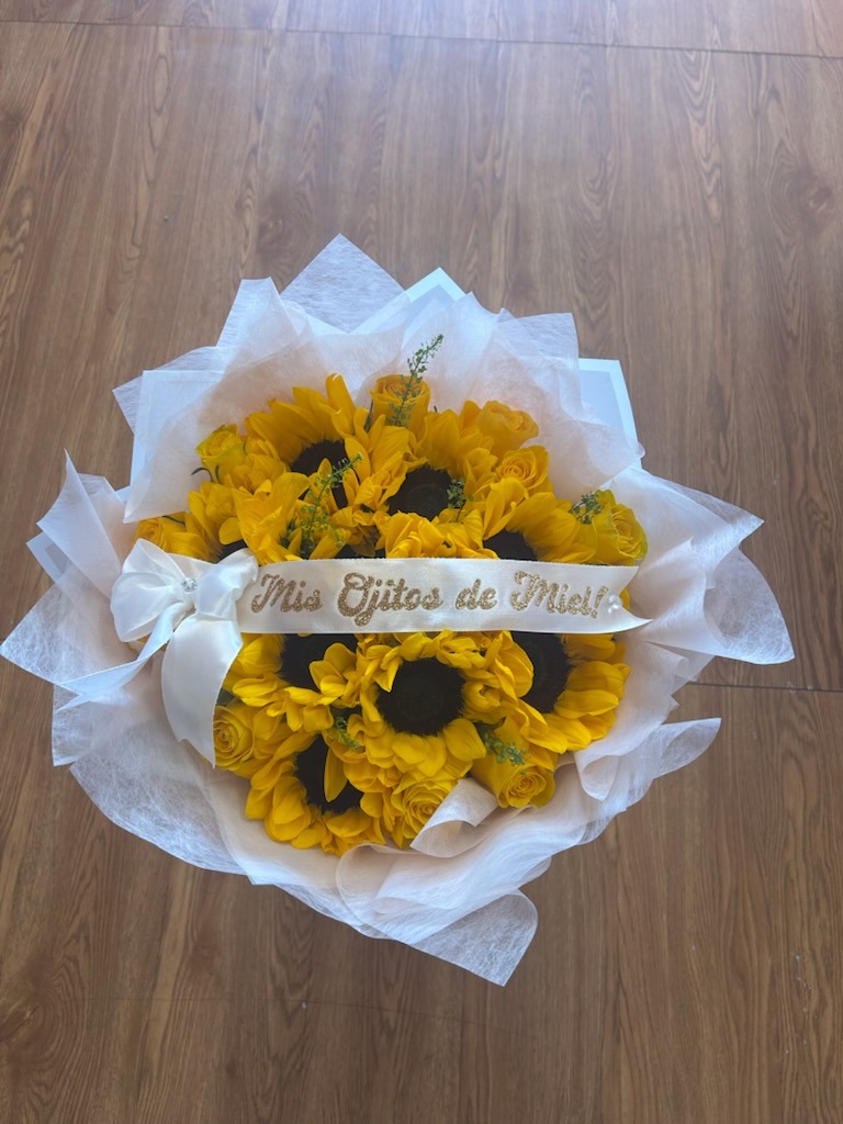 Vibrant sunflower and yellow Ecuadorian rose bouquet with custom 'Mis Ojitos de Miel!' glitter ribbon and white bow in delicate white wrap by FridAndre Florals