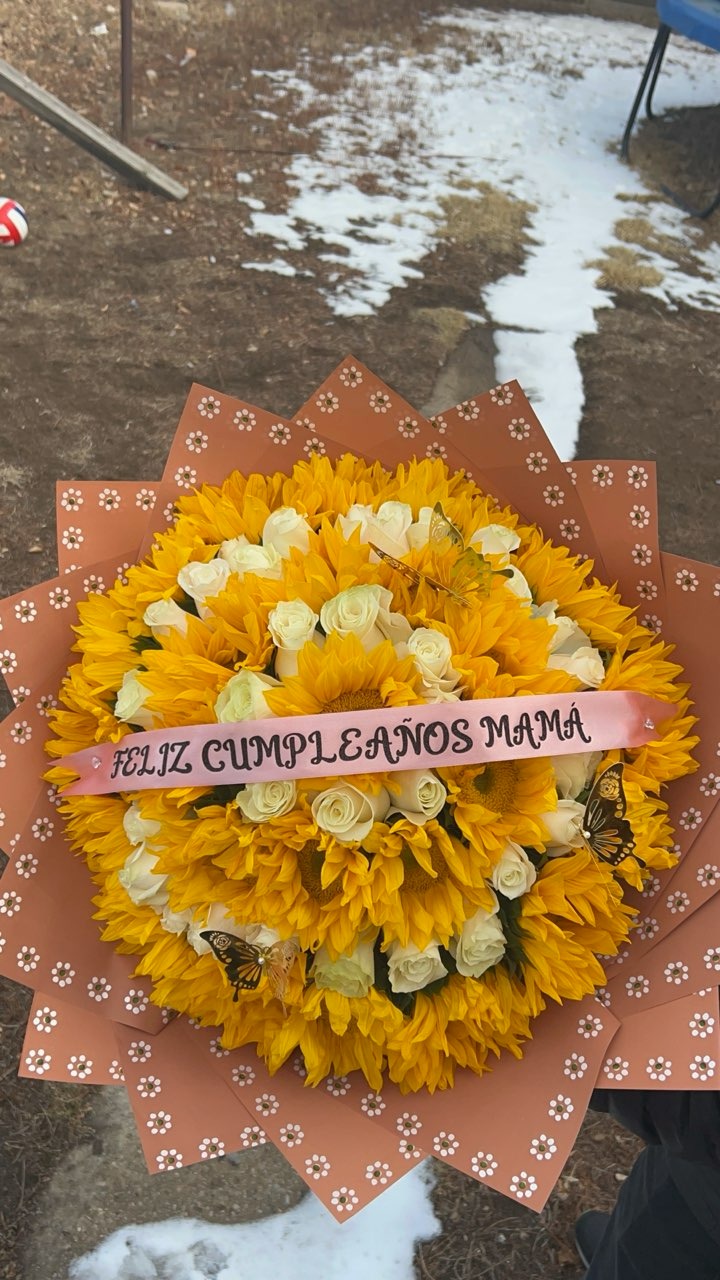Grand concentric sunflower and white Ecuadorian rose bouquet with 'FELIZ CUMPLEAÑOS MAMÁ' ribbon and butterfly accents in decorative star-point wrap by FridAndre Florals