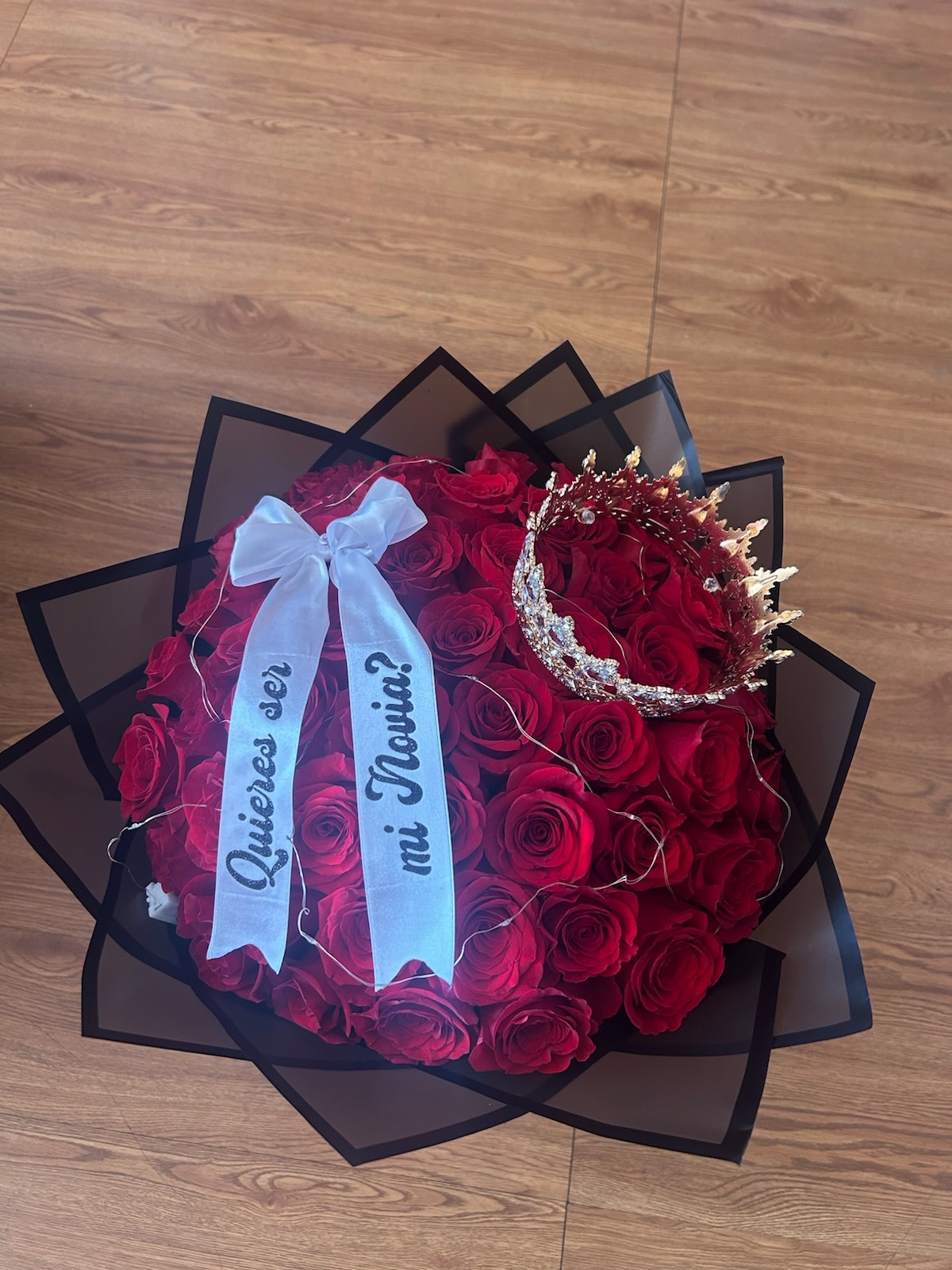 Romantic '¿Quieres ser mi Novia?' 50-rose deep red Ecuadorian rose bouquet with sparkling crystal crown tiara, twinkling LED fairy lights woven throughout blooms, '¿Quieres ser mi Novia?' silver script white satin ribbon bow, and elegant black geometric paper wrap - luxury illuminated proposal arrangement by FridAndre Florals