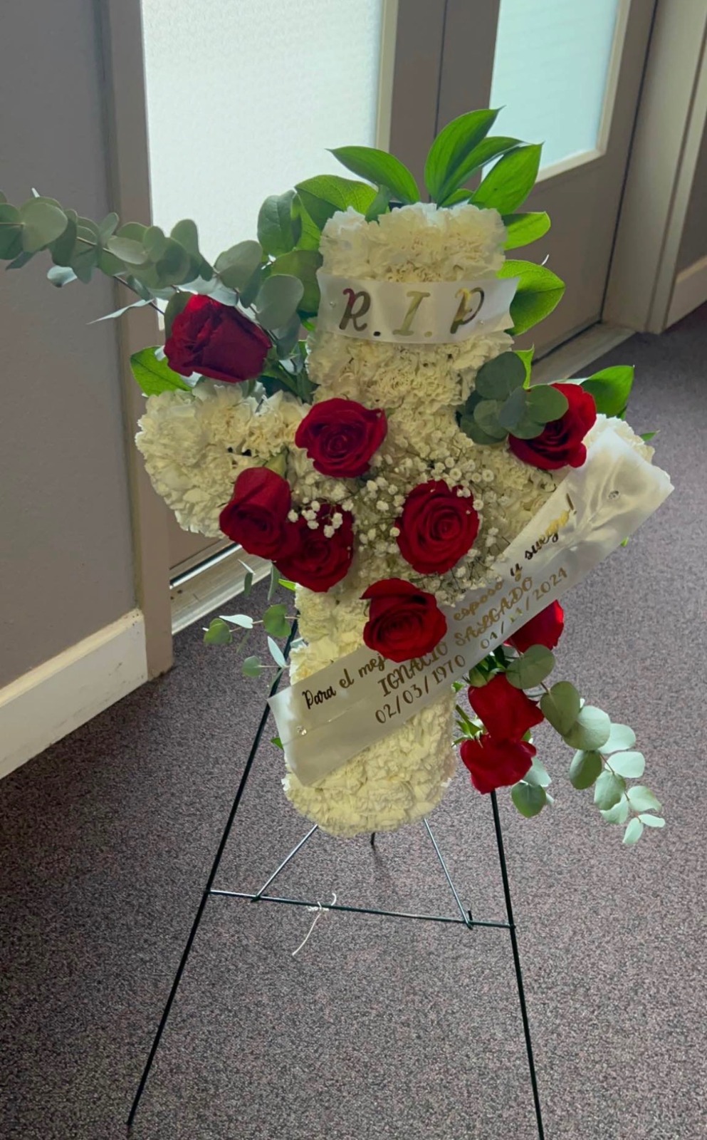 Small standing floral cross with white carnations, red Ecuadorian roses, fresh green eucalyptus leaves, 'R.I.P.' top ribbon, and 'Para el mejor IGNACIO SAGACIO 02/03/1970' memorial ribbon - small size sympathy arrangement