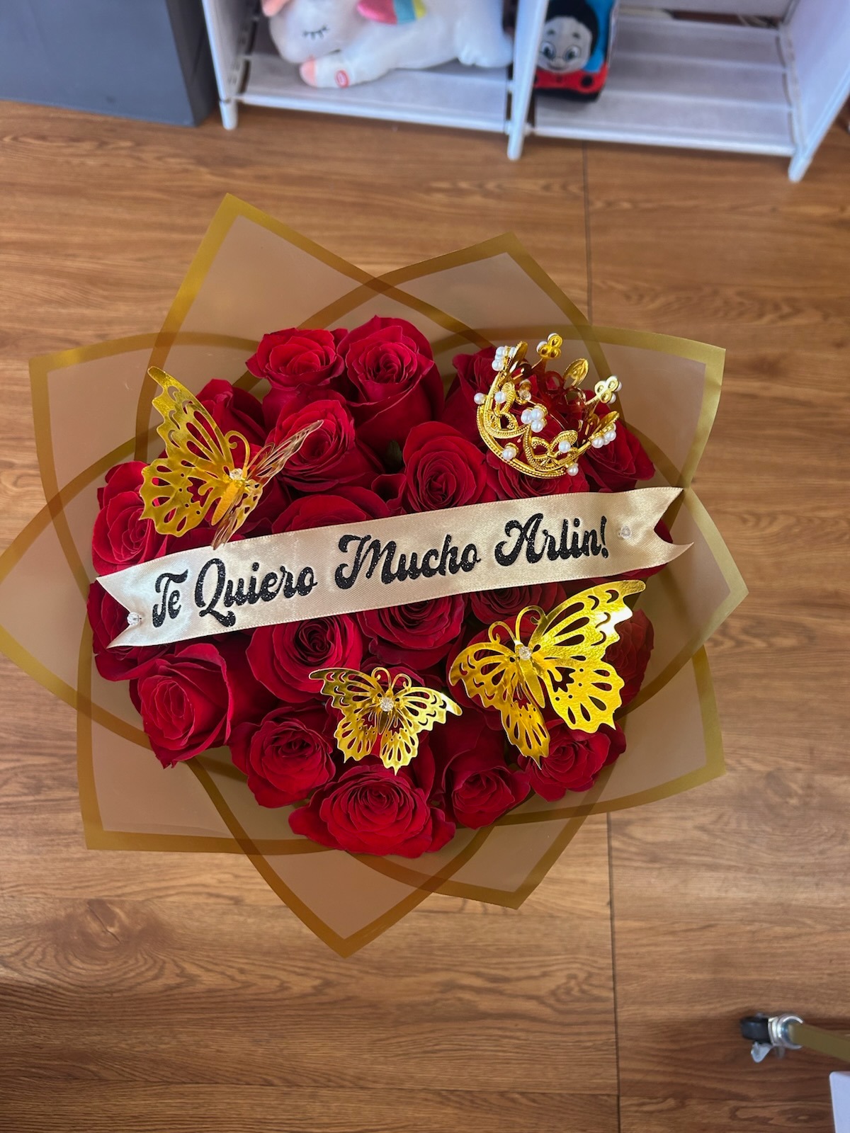 Romantic 'Te Quiero Mucho Arlin!' 25-rose deep red Ecuadorian round rose bouquet with sparkling crystal crown tiara with pearl accents, three golden laser-cut butterfly decorations, 'Te Quiero Mucho Arlin!' gold-glitter lettered cream satin ribbon bow, and elegant gold geometric paper wrap - love declaration arrangement by FridAndre Florals