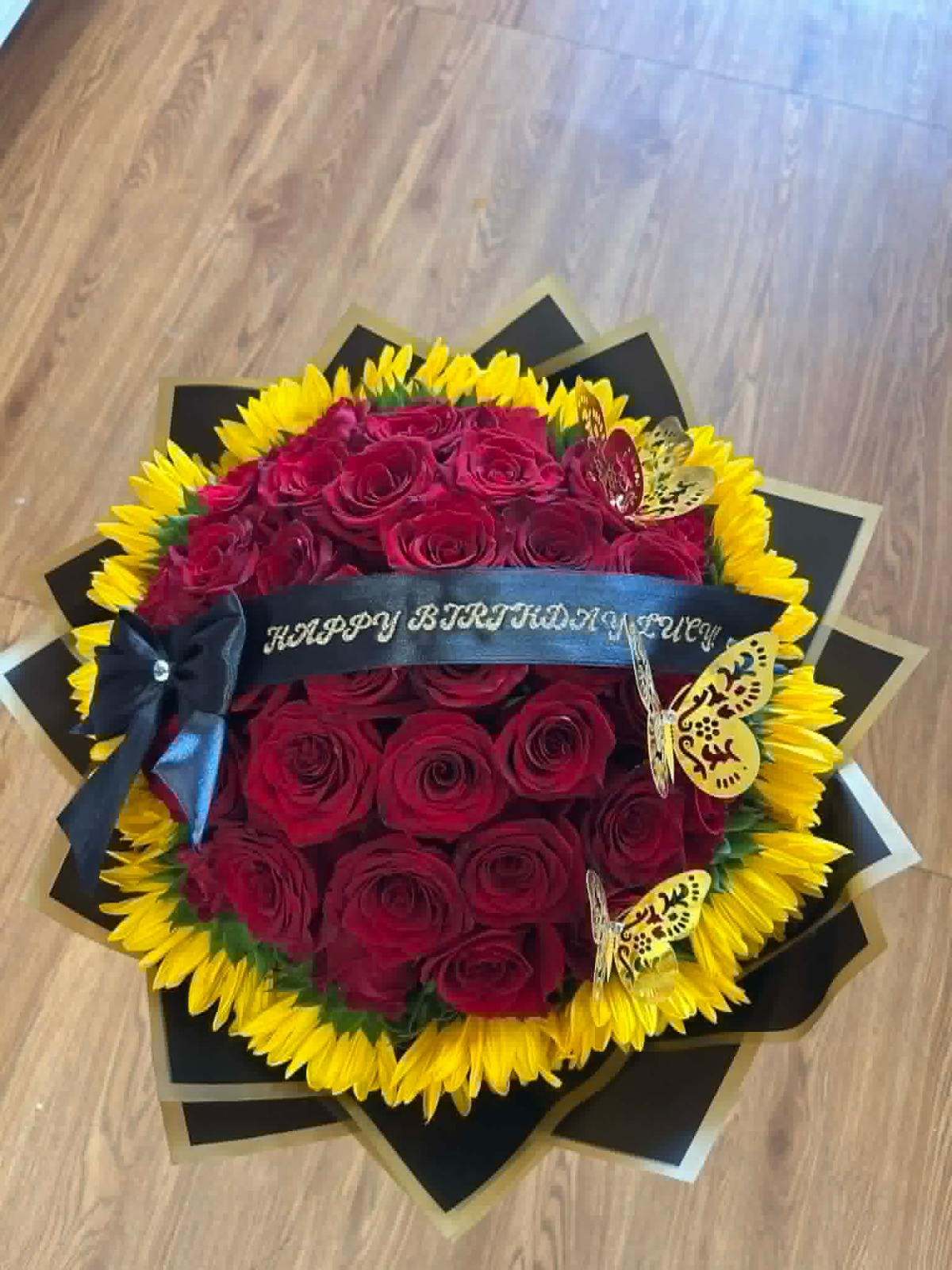 Stunning 'Happy Birthday Lucy!' 50-rose deep red Ecuadorian rose round bouquet surrounded by bright yellow sunflowers, golden laser-cut butterfly decorations, 'Happy Birthday Lucy' silver script black satin ribbon bow, sleek black geometric paper wrap with gold trim edges - luxury birthday celebration arrangement by FridAndre Florals