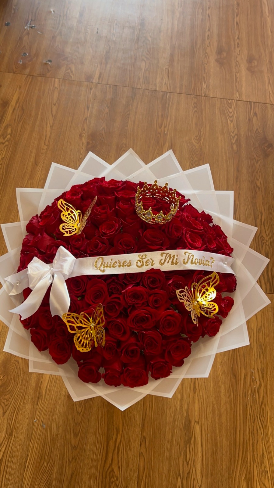 Stunning '¿Quieres Ser Mi Novia?' 75-rose deep red Ecuadorian round rose bouquet with sparkling crystal crown tiara, three golden laser-cut butterfly decorations, '¿Quieres Ser Mi Novia?' gold-glitter lettered white satin ribbon bow, elegant white geometric paper wrap, and large white satin ribbon accent - luxury proposal arrangement by FridAndre Florals