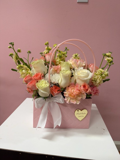 Quilted pink gift tote arrangement with white Ecuadorian roses, coral spray roses, peach carnations, snapdragons, white satin bow, and gold FridAndre heart charm