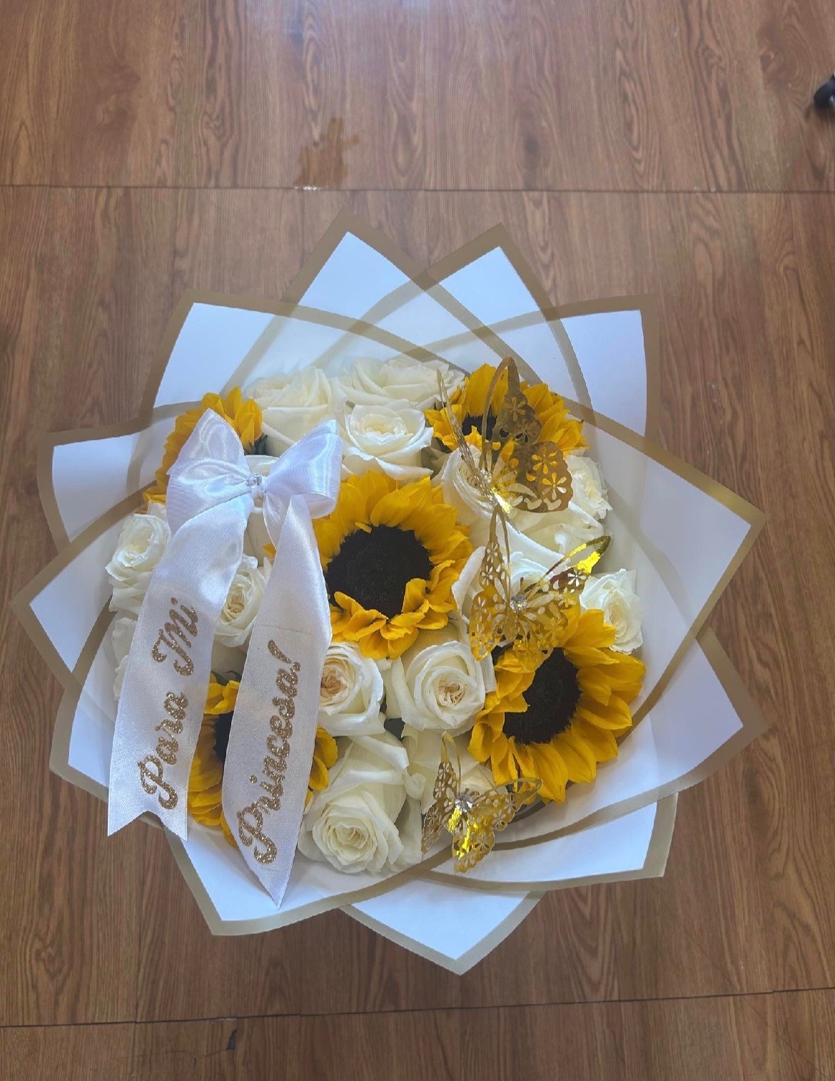 Beautiful white Ecuadorian rose bouquet with bright yellow sunflowers, golden laser-cut butterfly decorations, 'Para Ti Princesa!' gold-glitter lettered white satin ribbon bow, elegant white geometric paper wrap - cheerful princess gift arrangement