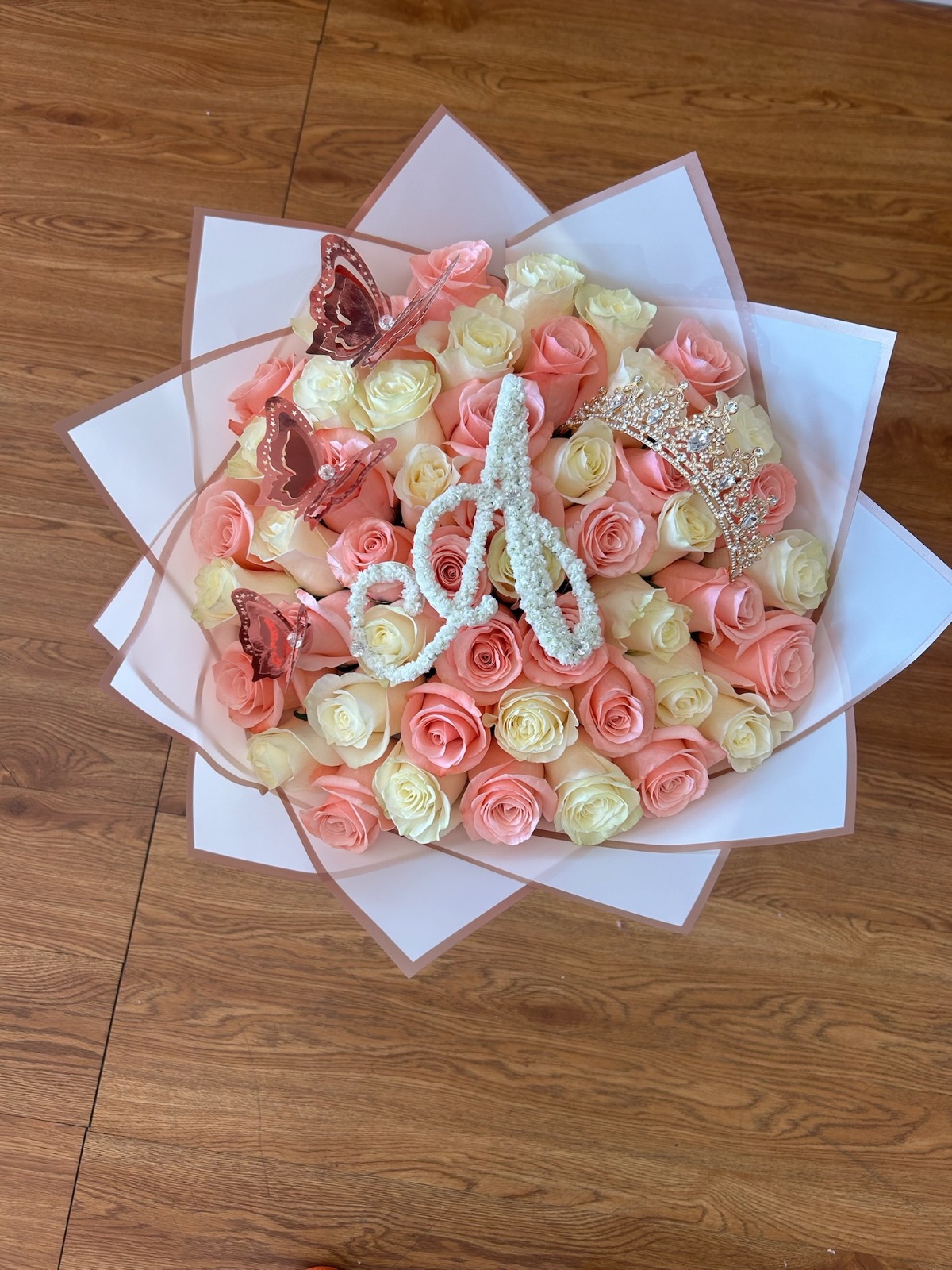 Stunning 50-rose pink and cream white Ecuadorian rose bouquet with sparkling crystal crown tiara, rose-gold laser-cut butterfly decorations, elegant cursive letter 'A' crafted from white baby's breath, and clean white geometric paper wrap - luxury personalized romantic arrangement by FridAndre Florals