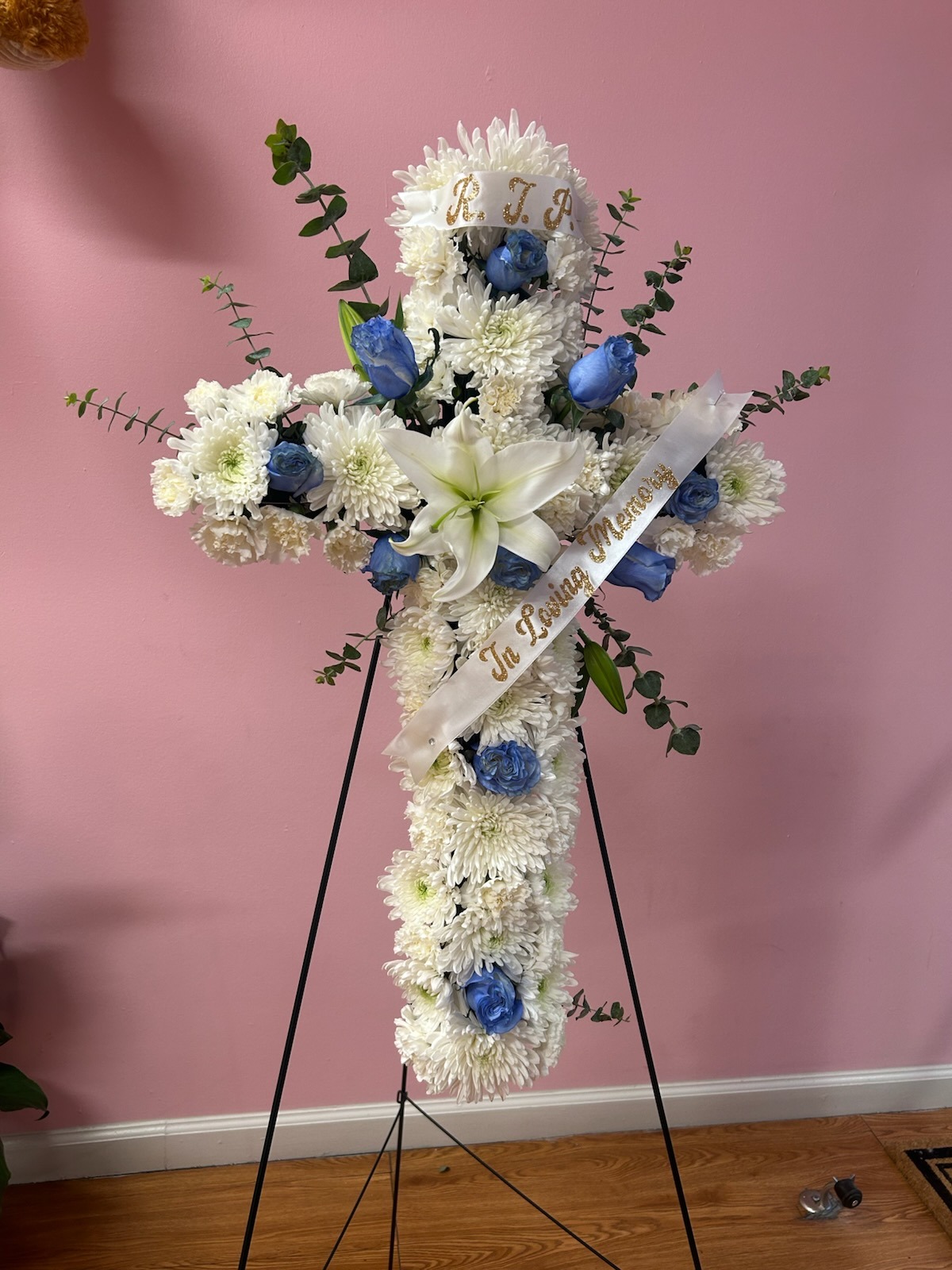 Elegant standing floral cross with white chrysanthemums, blue roses, white lily center, fresh green eucalyptus, 'R.I.P.' top ribbon, and 'In Loving Memory' diagonal memorial ribbon - medium size sympathy arrangement