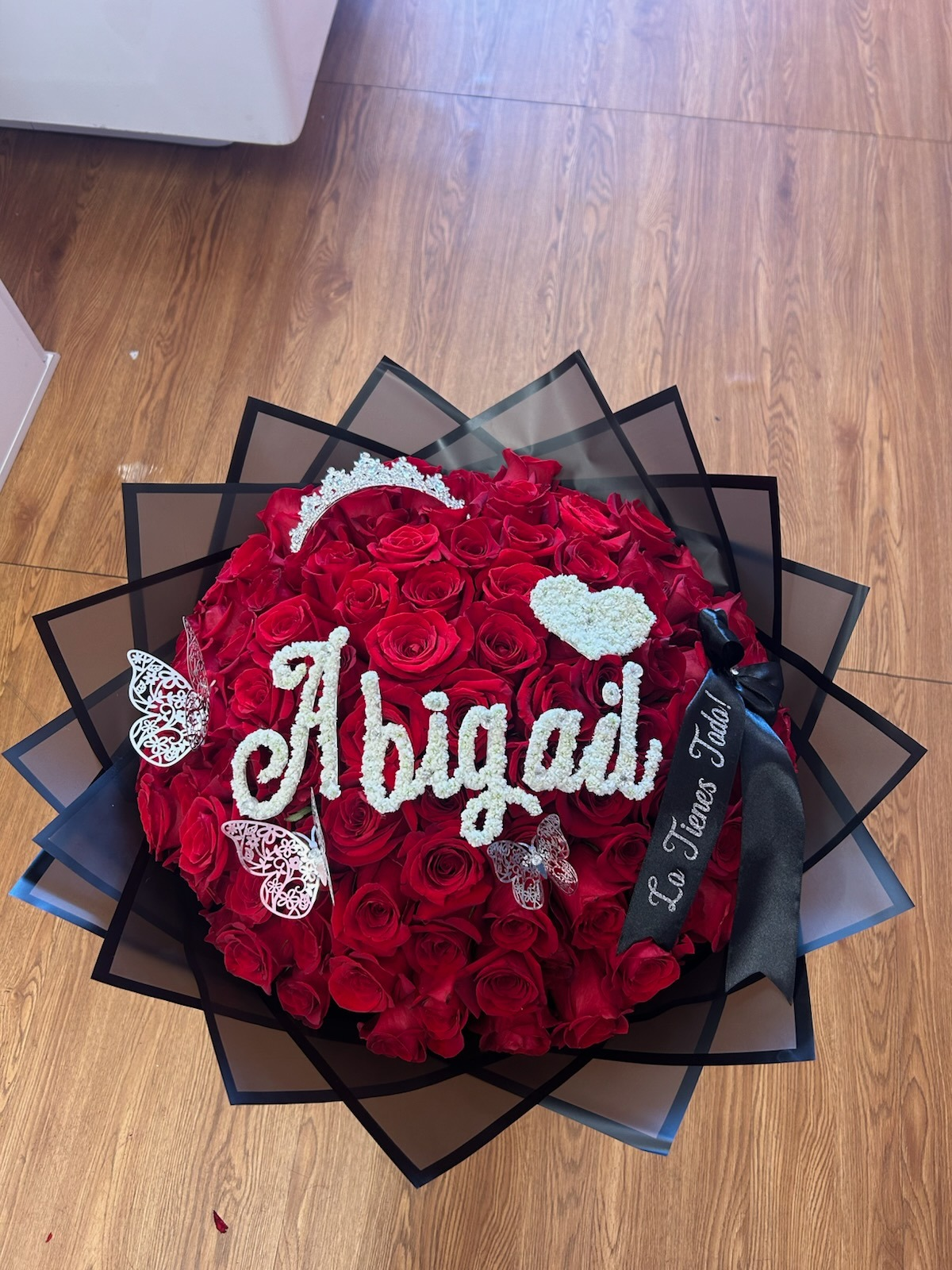 Stunning 'Abigail La Tienes Toda!' 100-rose deep red Ecuadorian round rose bouquet with sparkling crystal crown tiara, 'Abigail' name spelled in white baby's breath letters across blooms, small baby's breath heart accent, two golden laser-cut butterfly decorations, 'La Tienes Toda!' silver script black satin ribbon bow, and sleek black geometric paper wrap - luxury personalized romantic arrangement by FridAndre Florals