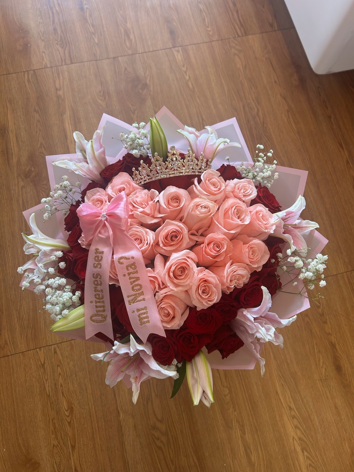 Stunning heart-shaped 50-rose pink and deep red Ecuadorian rose bouquet with sparkling crystal crown tiara, white lily accents, delicate white baby's breath border, '¿Quieres ser mi Novia?' gold-glitter lettered pink satin ribbon bow, and elegant pink geometric paper wrap - luxury proposal arrangement by FridAndre Florals