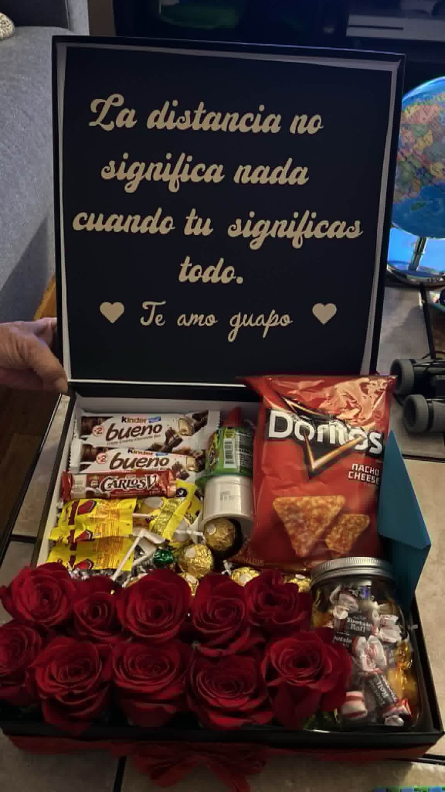 Romantic gift box with 12 red Ecuadorian roses, Doritos Nacho Cheese, Kinder Bueno, Carlos V chocolates, Ferrero Rocher, lollipops, and custom black lid engraved with 'La distancia no significa nada cuando tu significas todo. Te amo guapo' in gold lettering - long distance love gift arrangement