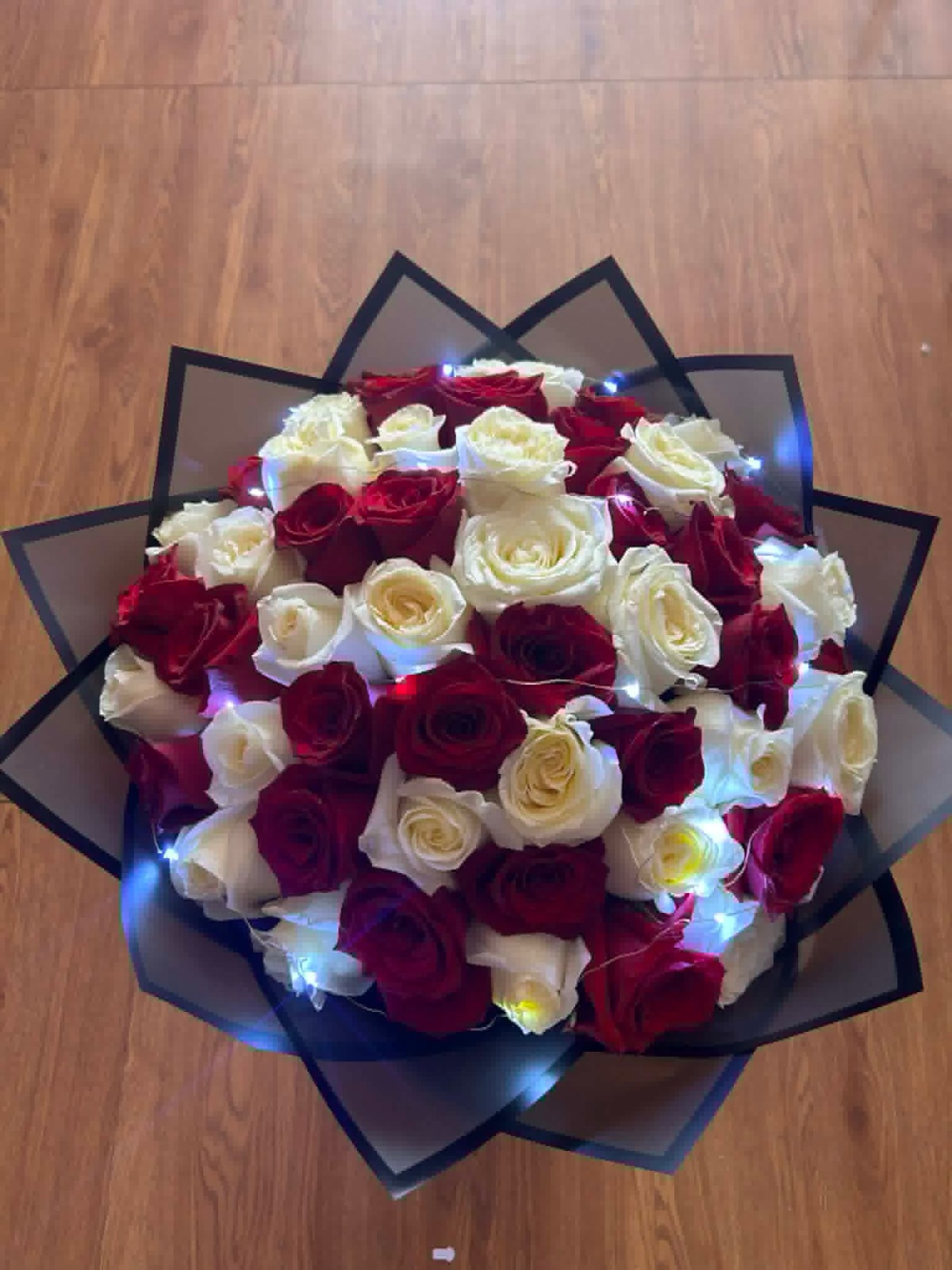 Stunning 50-rose red and cream white Ecuadorian rose round bouquet with twinkling LED fairy lights woven throughout blooms, elegant black geometric paper wrap with gold trim edges - luxury illuminated romantic arrangement by FridAndre Florals
