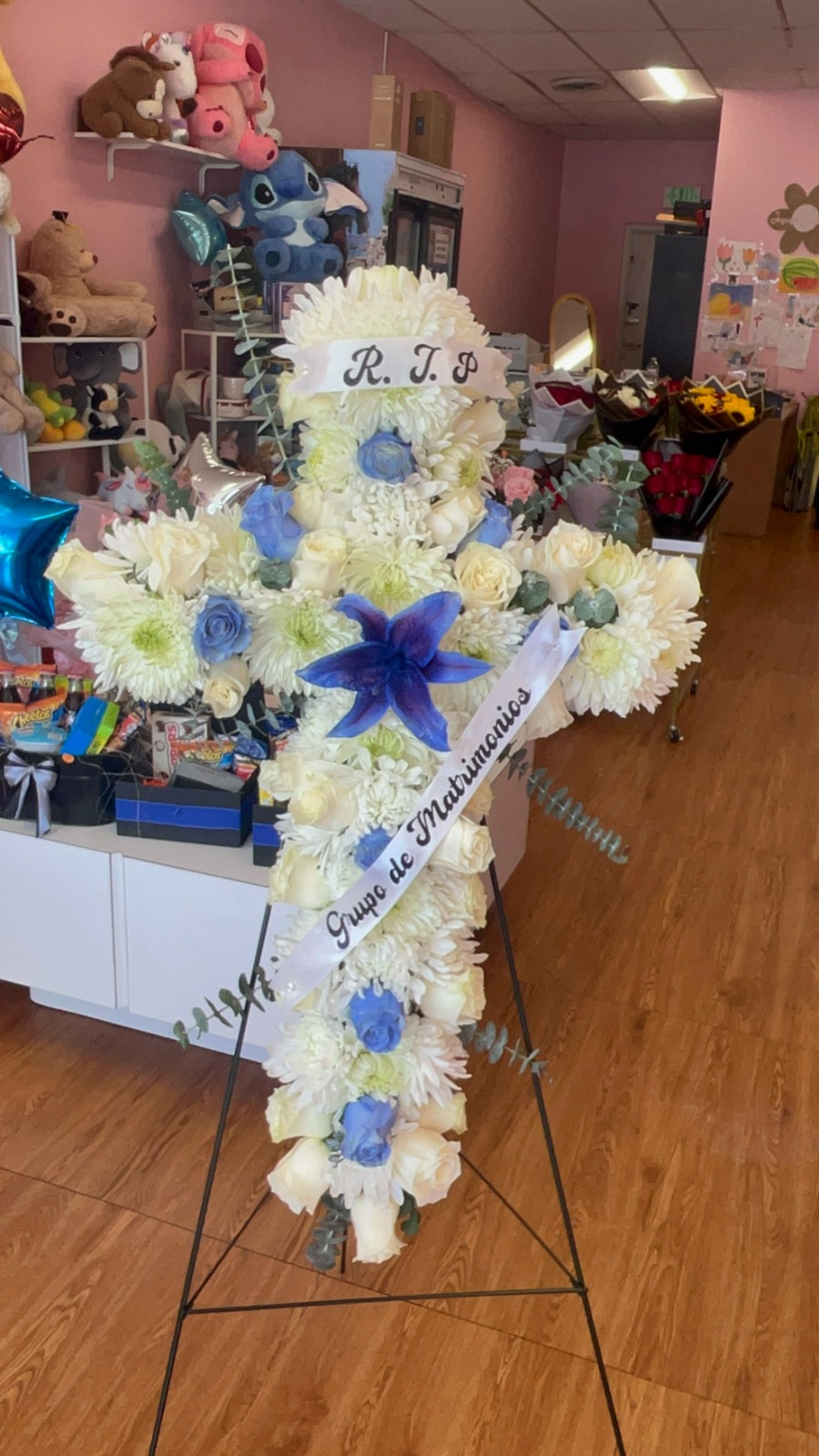 Elegant standing floral cross with white chrysanthemums, blue roses, white lily center, fresh green eucalyptus, 'R.I.P.' top ribbon, and 'Grupo de Matrimonios' memorial ribbon - medium size