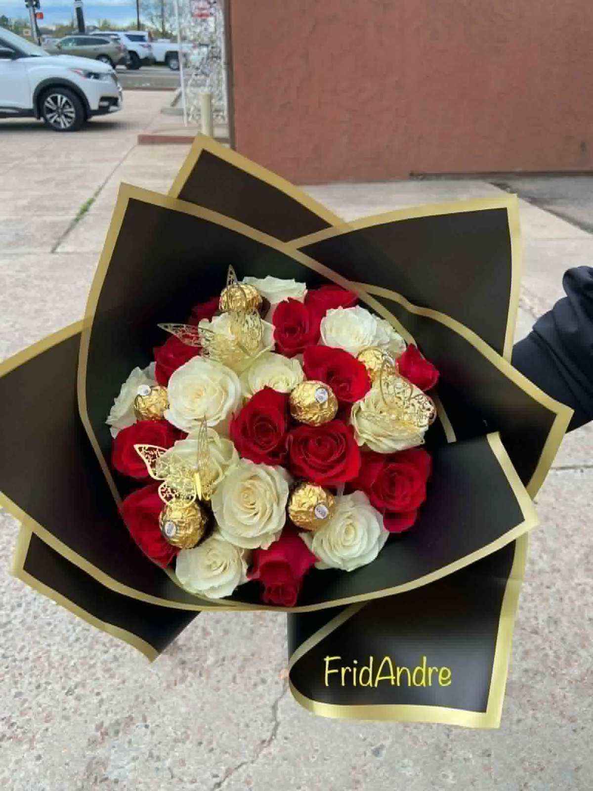 Luxury red and white Ecuadorian rose bouquet with 25 roses, 6 Ferrero Rocher chocolates nestled among blooms, golden laser-cut butterfly decorations, elegant black geometric paper wrap with gold trim edges, 'FridAndre' gold lettering on wrap - premium chocolate rose gift arrangement