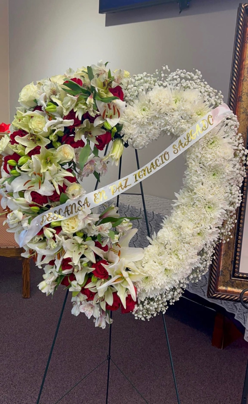 Large heart-shaped sympathy standing wreath with white lilies, red Ecuadorian roses, white chrysanthemums, baby's breath border, and 'Descansa En Paz Ignacio Sagacio' gold-lettered memorial ribbon - medium size