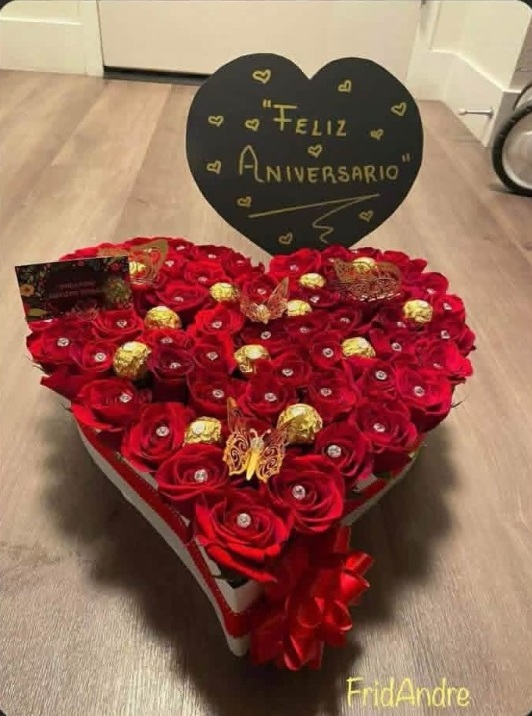 Luxury heart-shaped red rose gift box with 'Feliz Aniversario' anniversary sign, Ferrero Rocher chocolates nestled among blooms, golden laser-cut butterfly decorations, crystal rhinestone rose accents, and large red satin ribbon bow - medium size