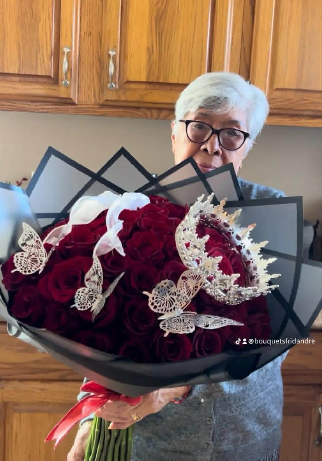Massive 50-rose deep red Ecuadorian rose bouquet with sparkling crystal crown tiara, golden laser-cut butterfly decorations, elegant black geometric paper wrap with gold trim edges, white calla lily accents, and vibrant red satin ribbon bow - luxury premium romantic arrangement