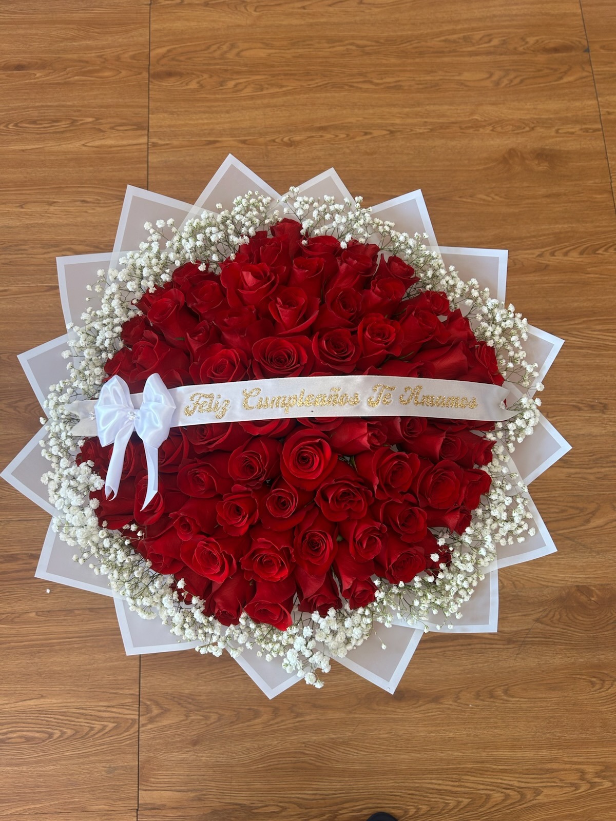 Stunning heart-shaped 75-rose deep red Ecuadorian rose bouquet with white baby's breath border, 'Feliz Cumpleaños Te Amamos' gold-glitter lettered white satin ribbon bow, and elegant white geometric paper wrap - luxury birthday love declaration arrangement by FridAndre Florals