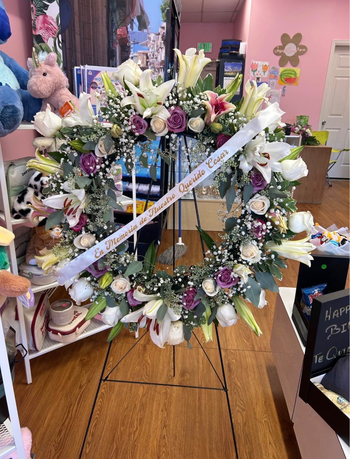 Heart-shaped sympathy standing wreath with white lilies, purple and cream Ecuadorian roses, delicate white baby's breath border, fresh green eucalyptus leaves, and 'En Memoria de Nuestro Querido Cesar' gold-lettered memorial ribbon - medium size dignified tribute arrangement