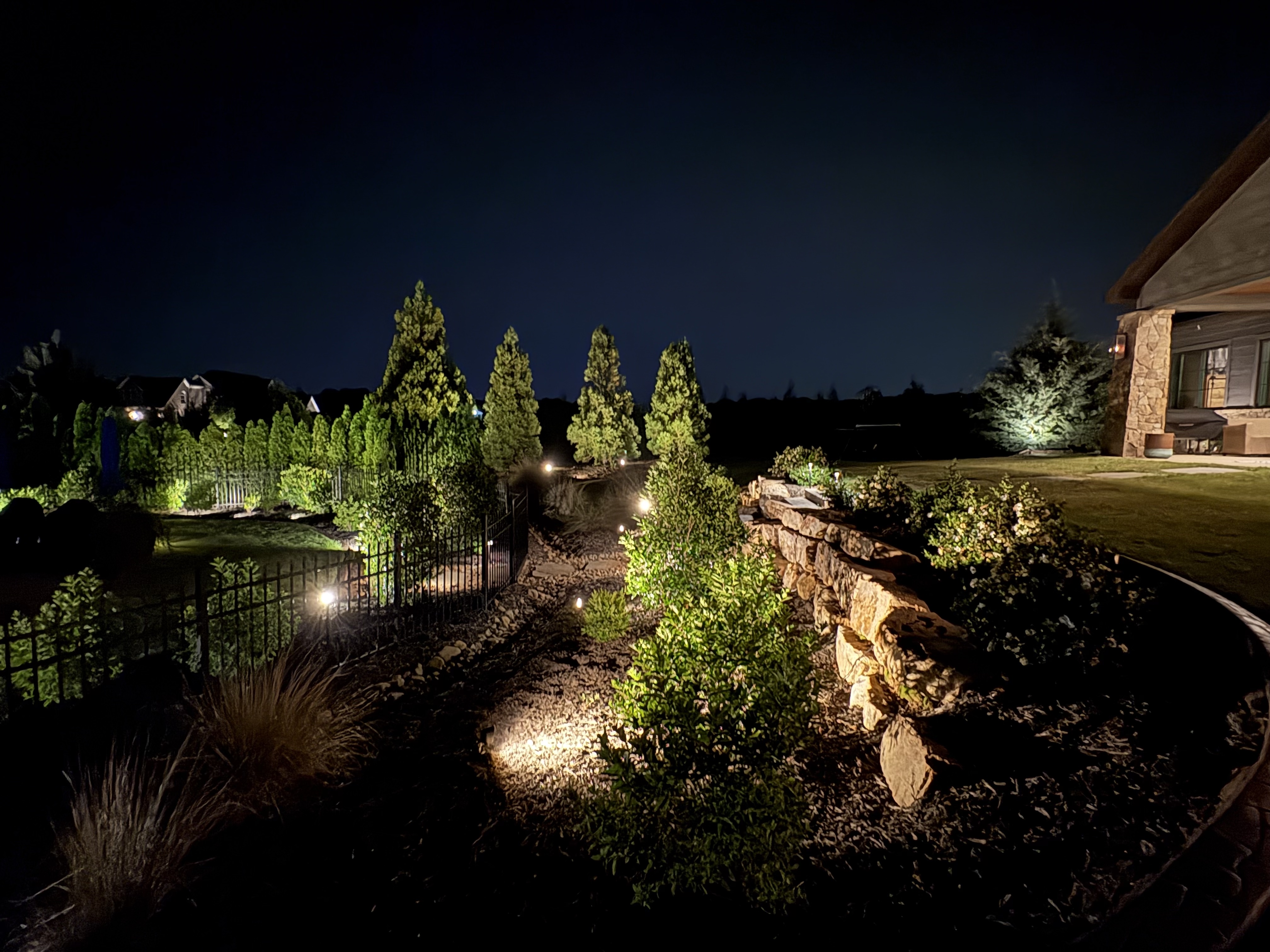 Backyard retaining wall path lighting with evergreen perimeter and illuminated stone terrace in Greenville SC