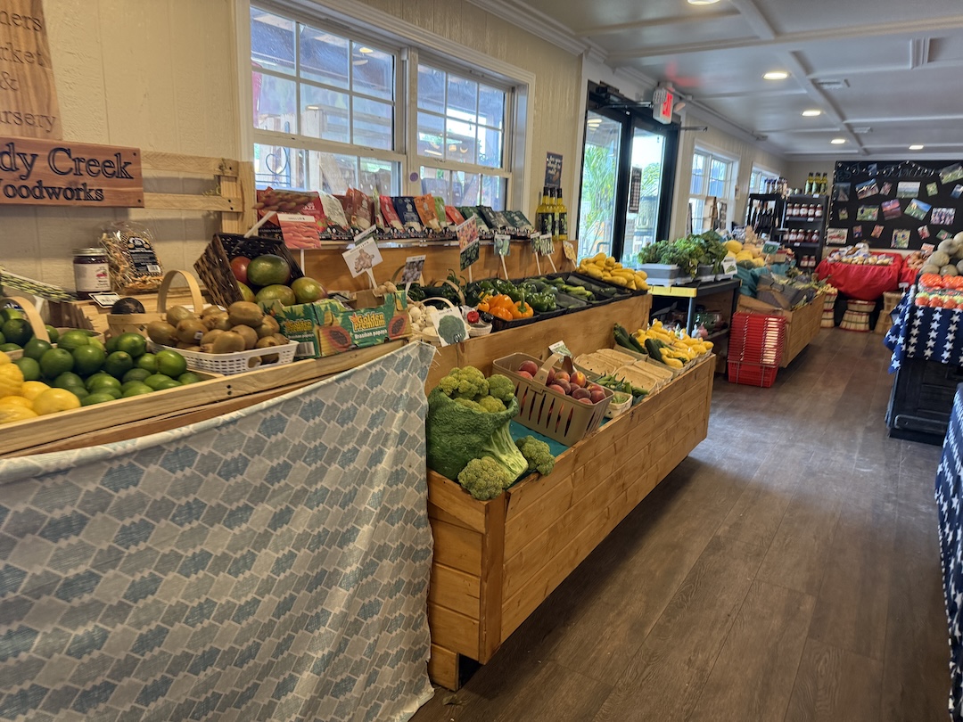 Interior of Hughes Farmers Market produce department showing wooden bins filled with fresh limes, lemons, kiwis, potatoes, broccoli, peaches, cucumbers, squash, peppers, and seasonal fruits under large windows