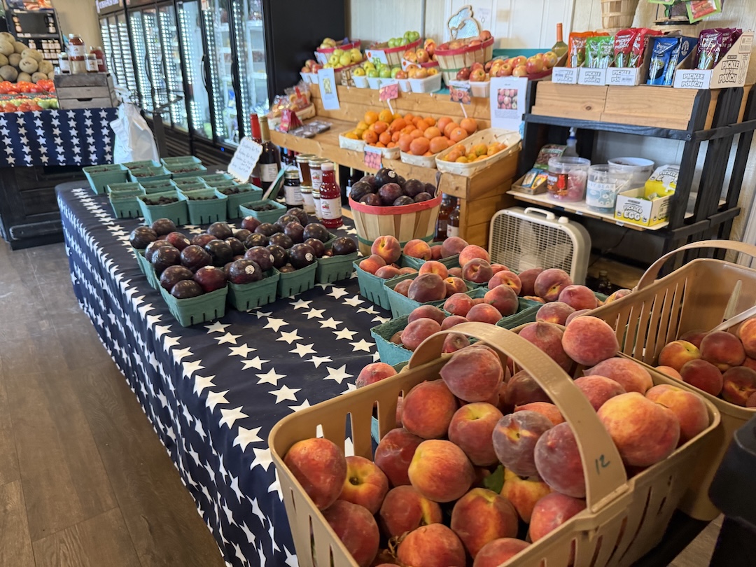 Hughes Farmers Market fruit table featuring baskets of ripe peaches, plums in green pint containers, oranges, apples, local honey, jams, jellies, and preserves on a patriotic star-patterned tablecloth
