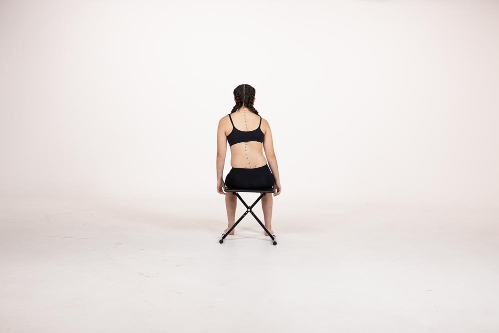 A woman with braided hair sitting on a stool with her back facing the camera, demonstrating a ScoliBalance corrective posture