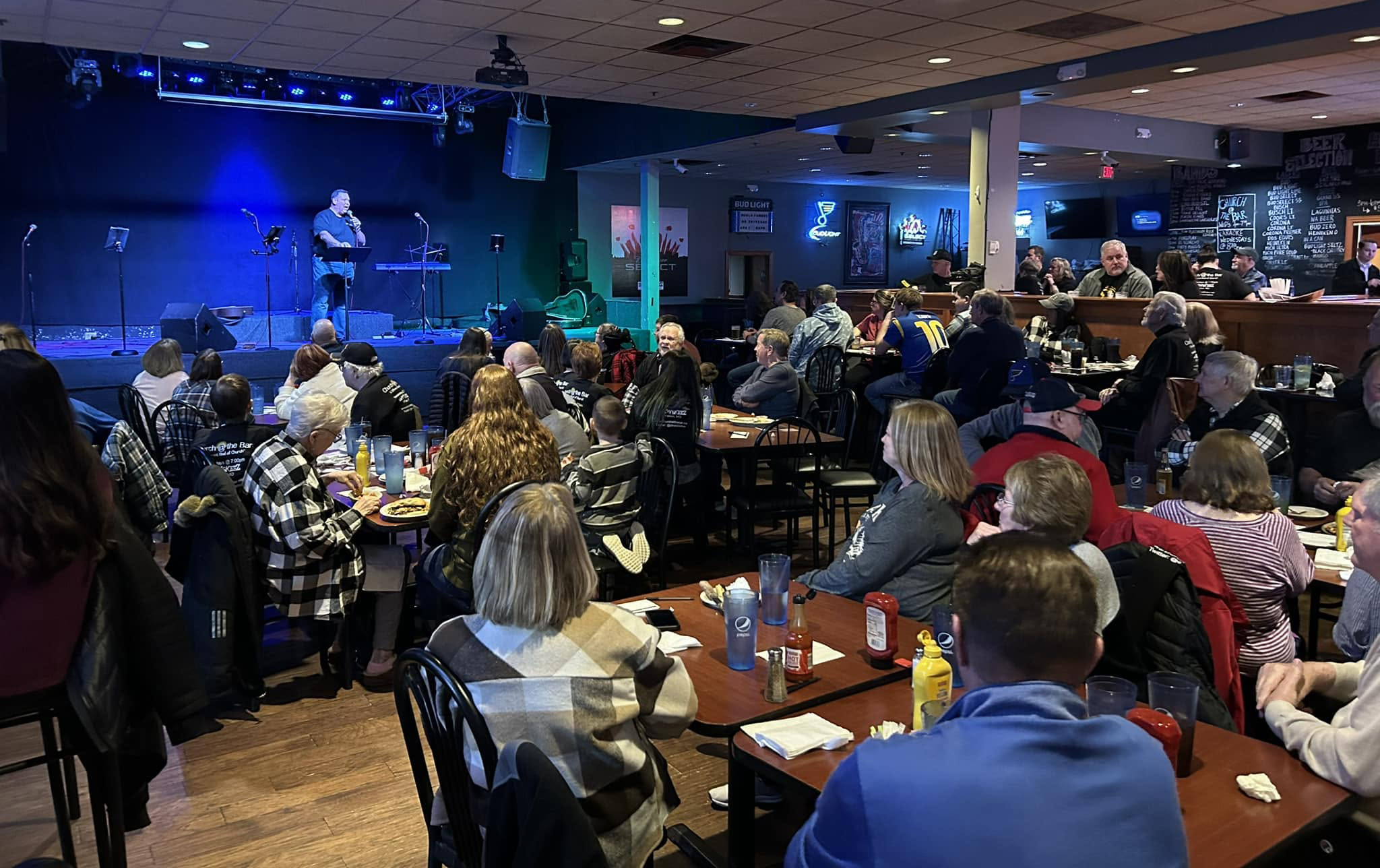 Church at the Bar service happening inside Brewskeez bar with people gathered at tables listening to a speaker on stage