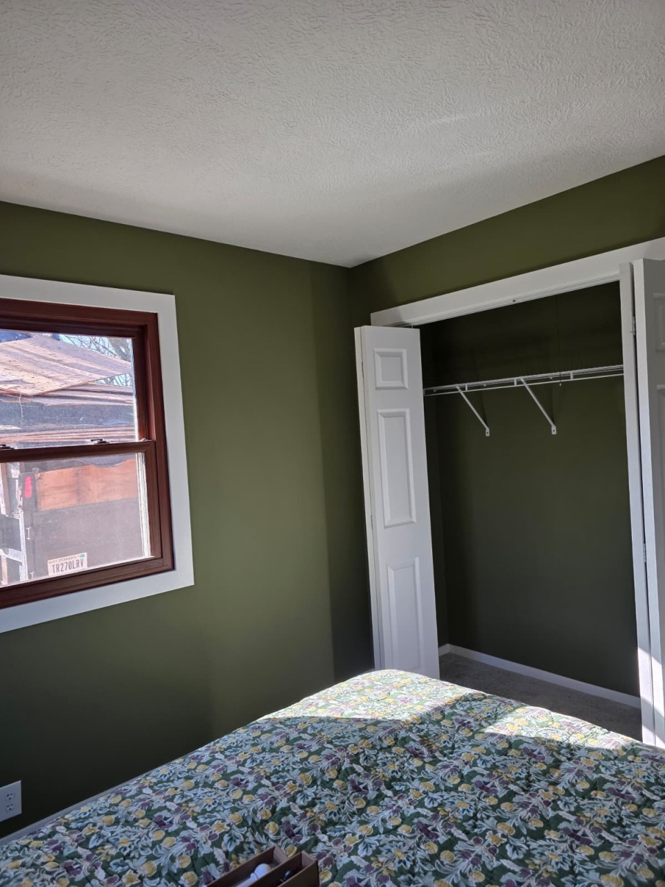 Olive green bedroom featuring window with wood trim, white closet doors, and freshly painted walls with smooth texture finish