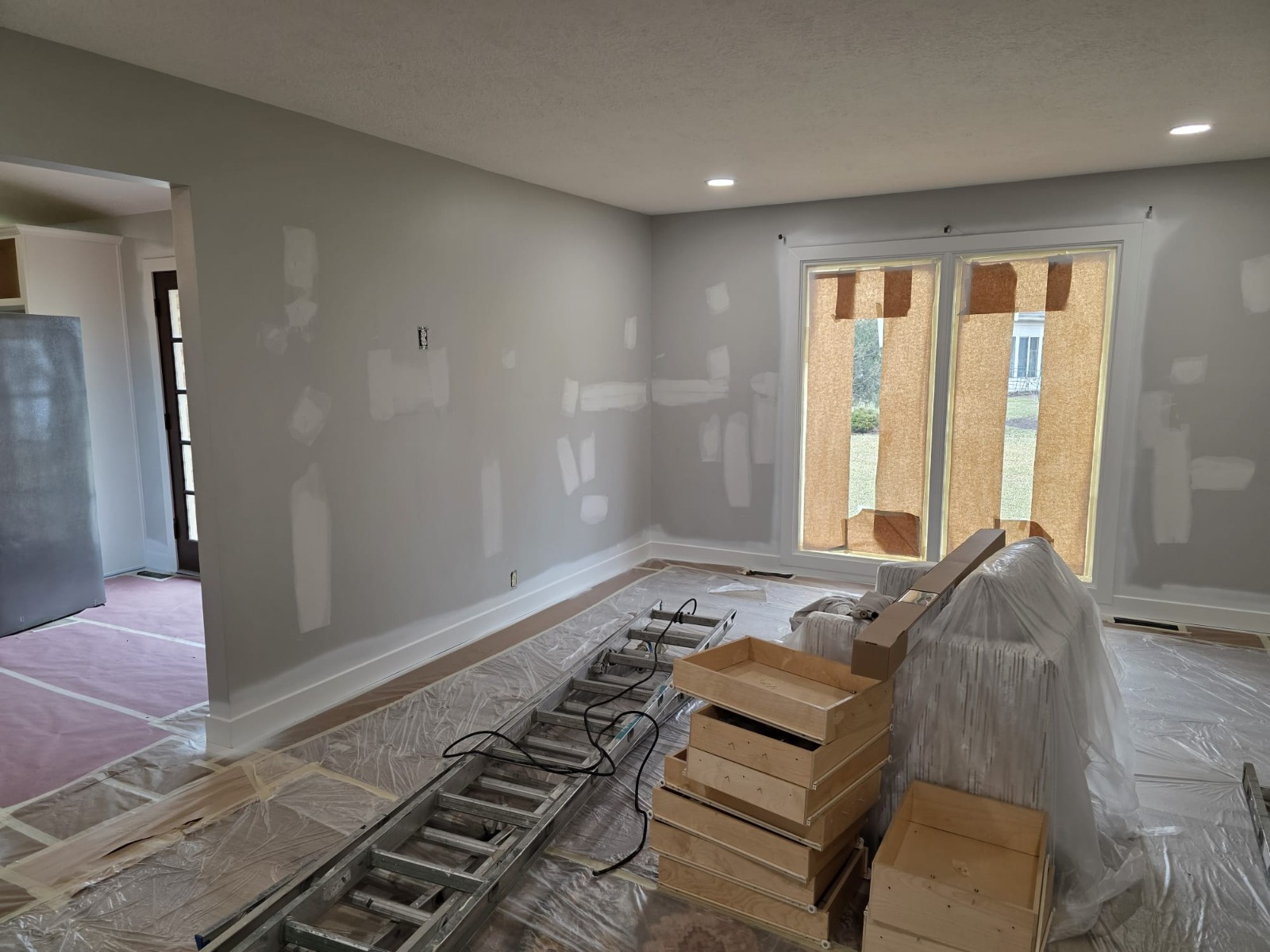 Living room under renovation with plastic sheeting protecting floors, furniture covered, and ladder ready for painting
