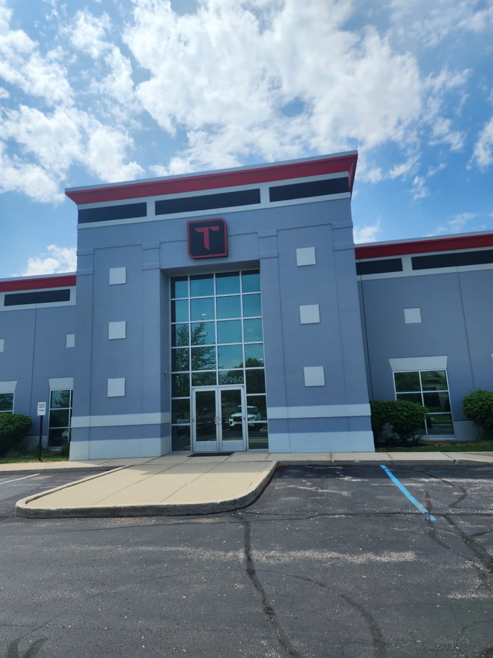 Commercial building entrance with gray facade, red roof trim, glass double doors, and prominent company logo above entrance