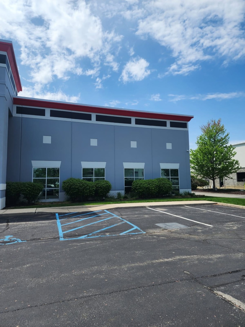 Commercial building exterior showing long gray wall with red trim along roofline, large windows, and landscaped parking area