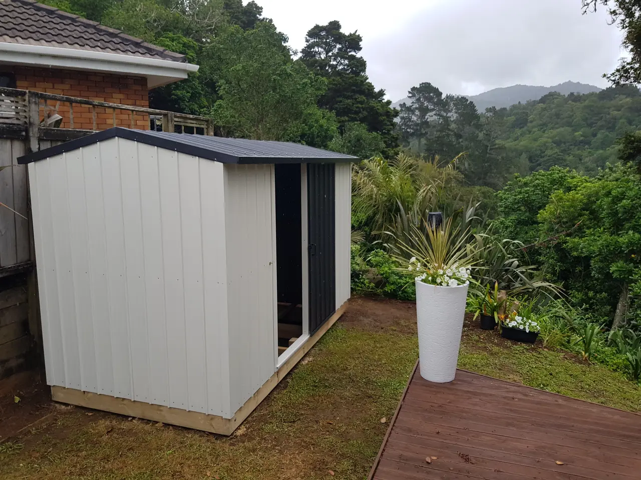 Garden Shed with Timber Decking - The completed garden shed with black roof among established trees, with a timber deck and planter giving the outbuilding a settled, lived-in feel.