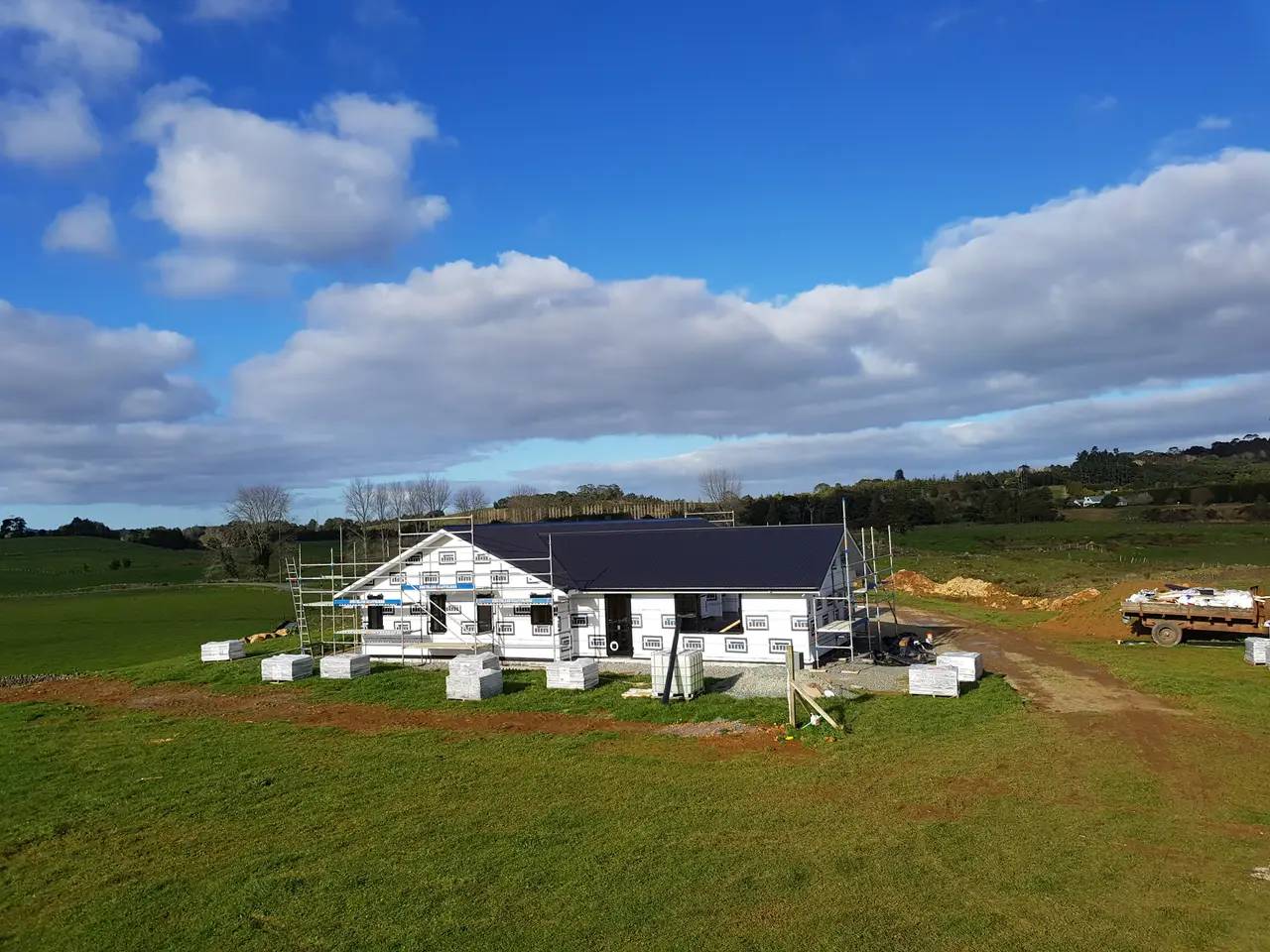 Rural Residential New Build & Roof Installation - Rural residential home build underway, with weathertight building wrap and dark long-run roof installed, scaffold access up and cladding materials staged on site.