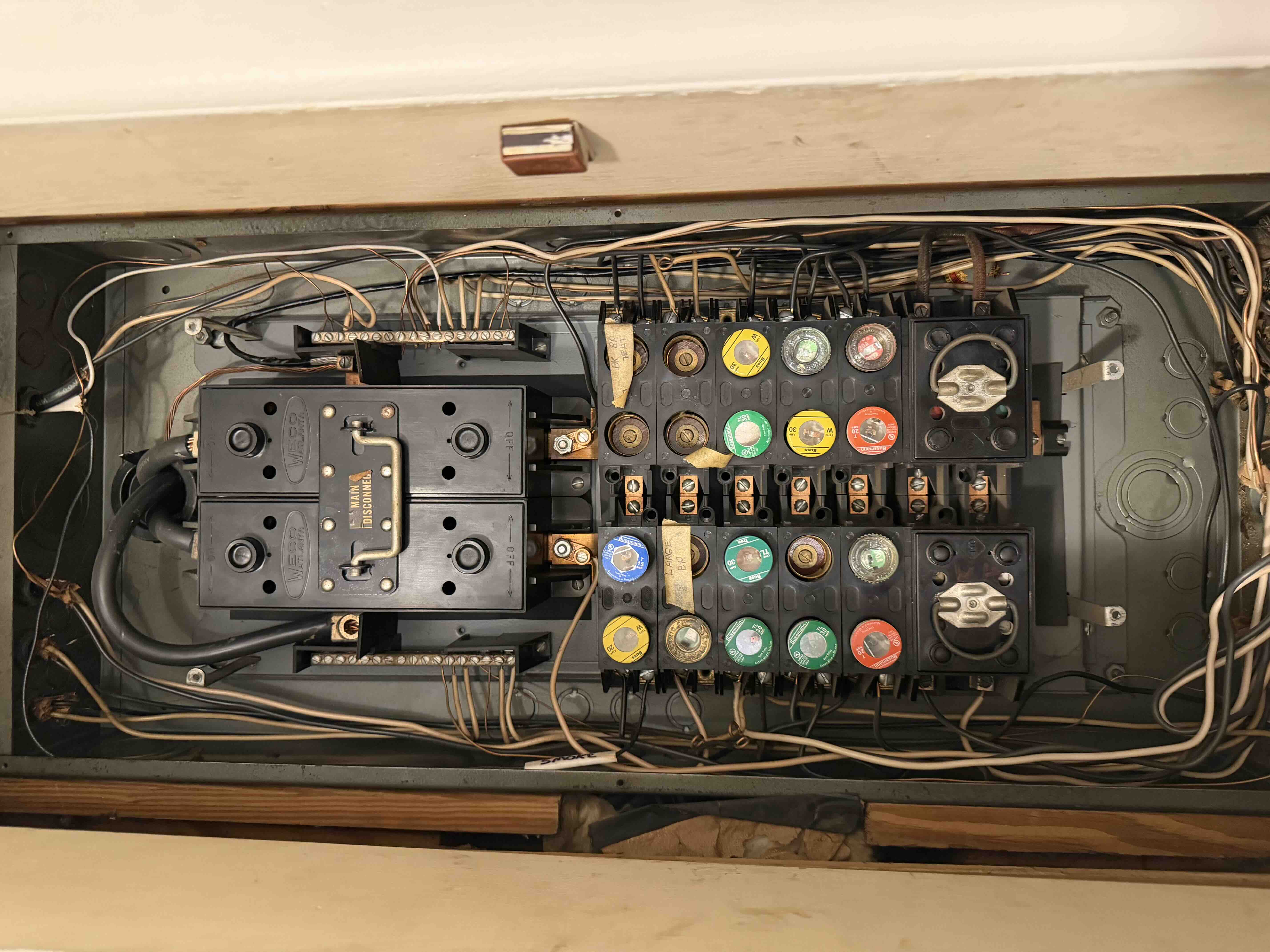 Old fuse panel interior with tangled wiring and aging fuse blocks prior to a breaker panel upgrade