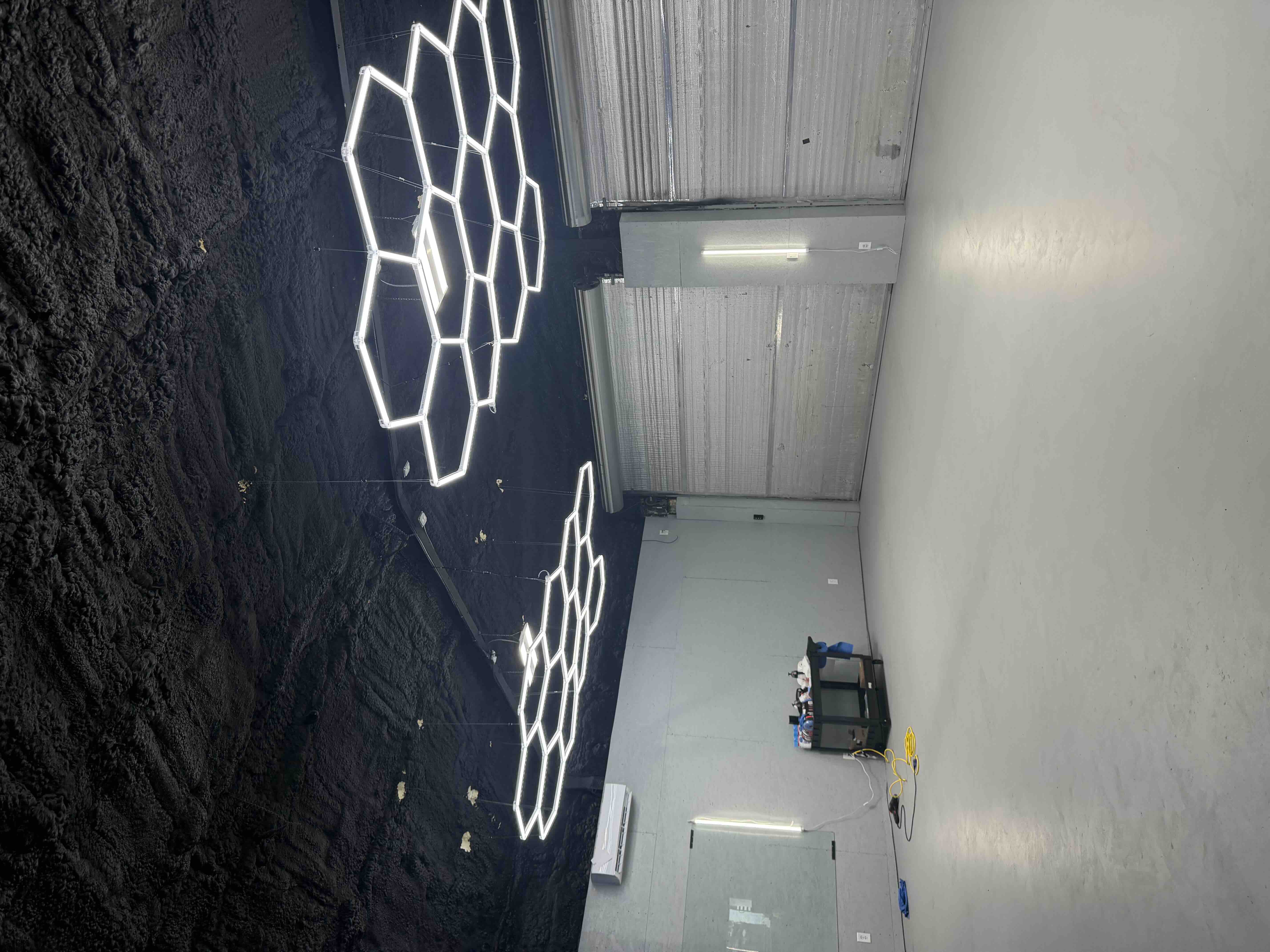 Two illuminated hexagon LED honeycomb light fixtures suspended by wires in a shop during fabrication