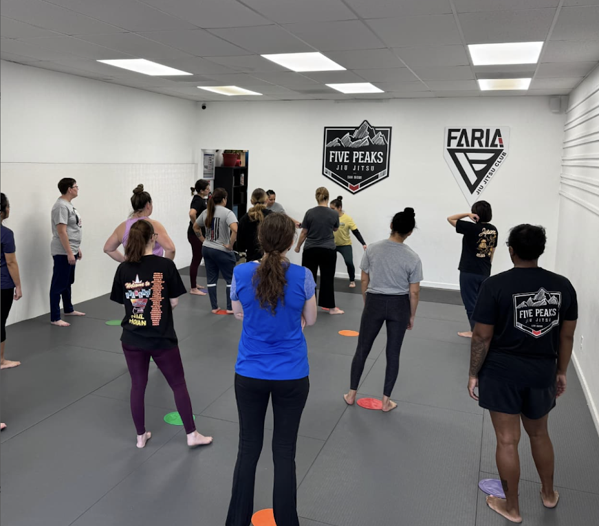 Students standing on the mat during a group warm-up class at Five Peaks Jiu Jitsu, with Five Peaks and Faria Jiu Jitsu Club logos on the wall