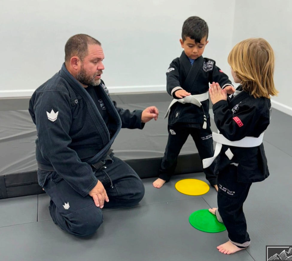 Coach kneeling with two young students practicing jiu jitsu technique on the mat at Five Peaks Jiu Jitsu