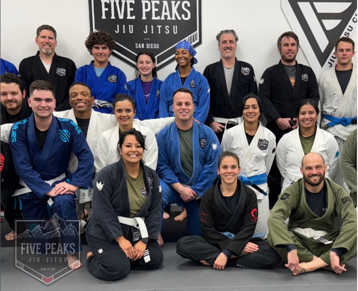 Coaches and adult students posing together on the mat in front of the Five Peaks Jiu Jitsu logo