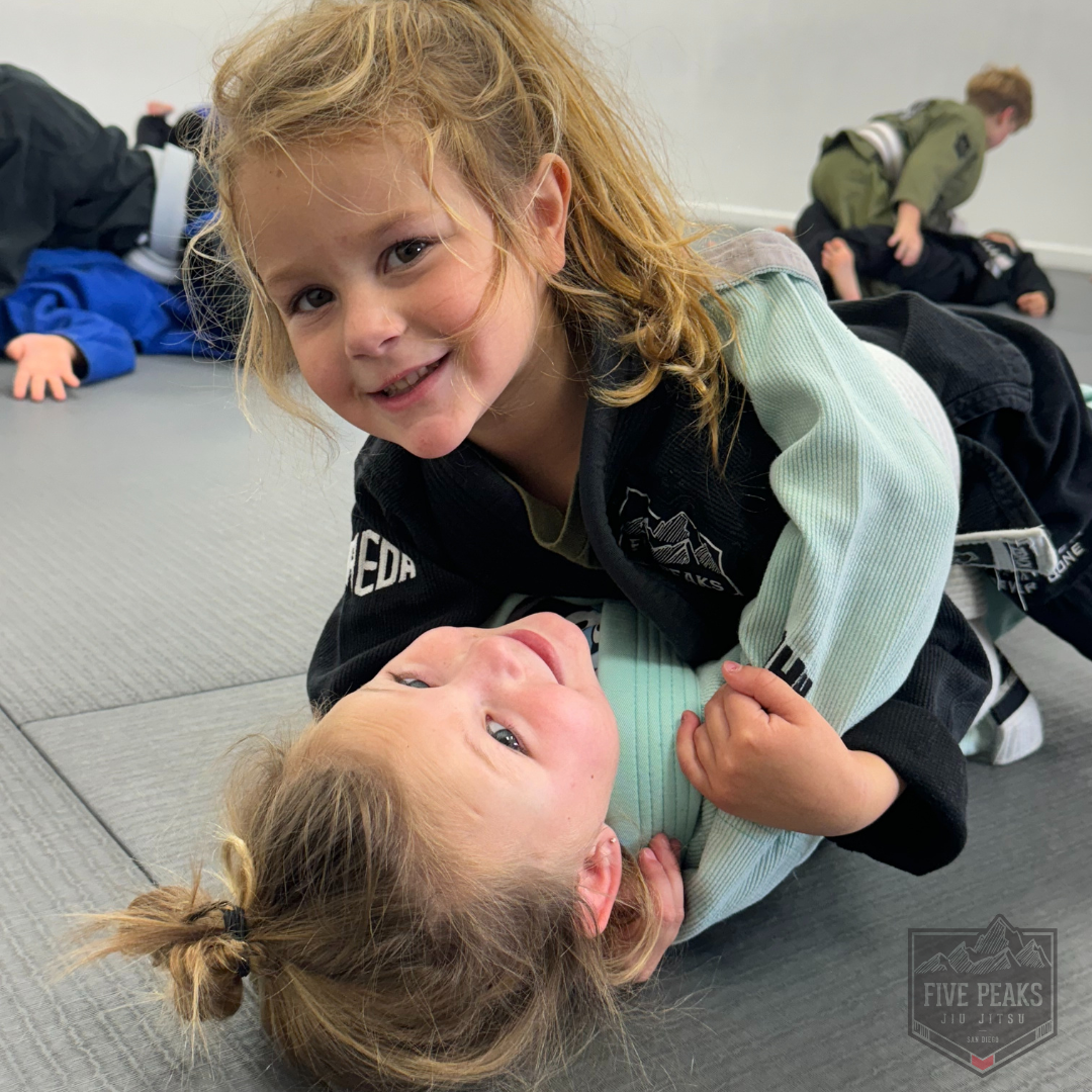 Two young girls smiling during jiu jitsu ground training at Five Peaks Jiu Jitsu