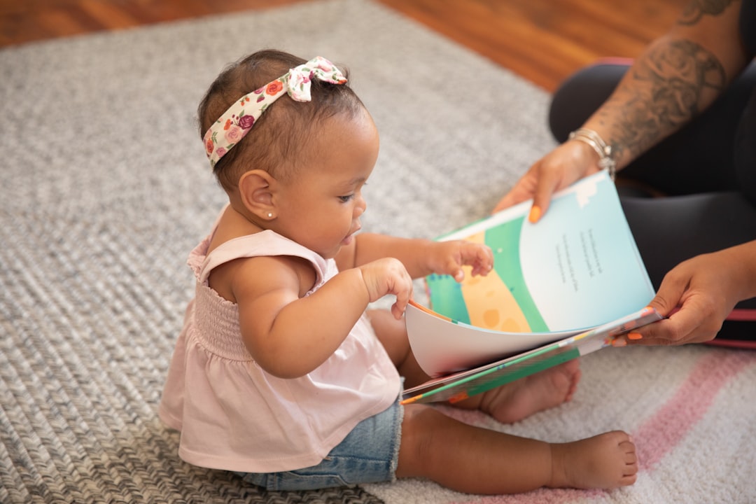 A baby sitting on a rug looking at a picture book held by an adult.