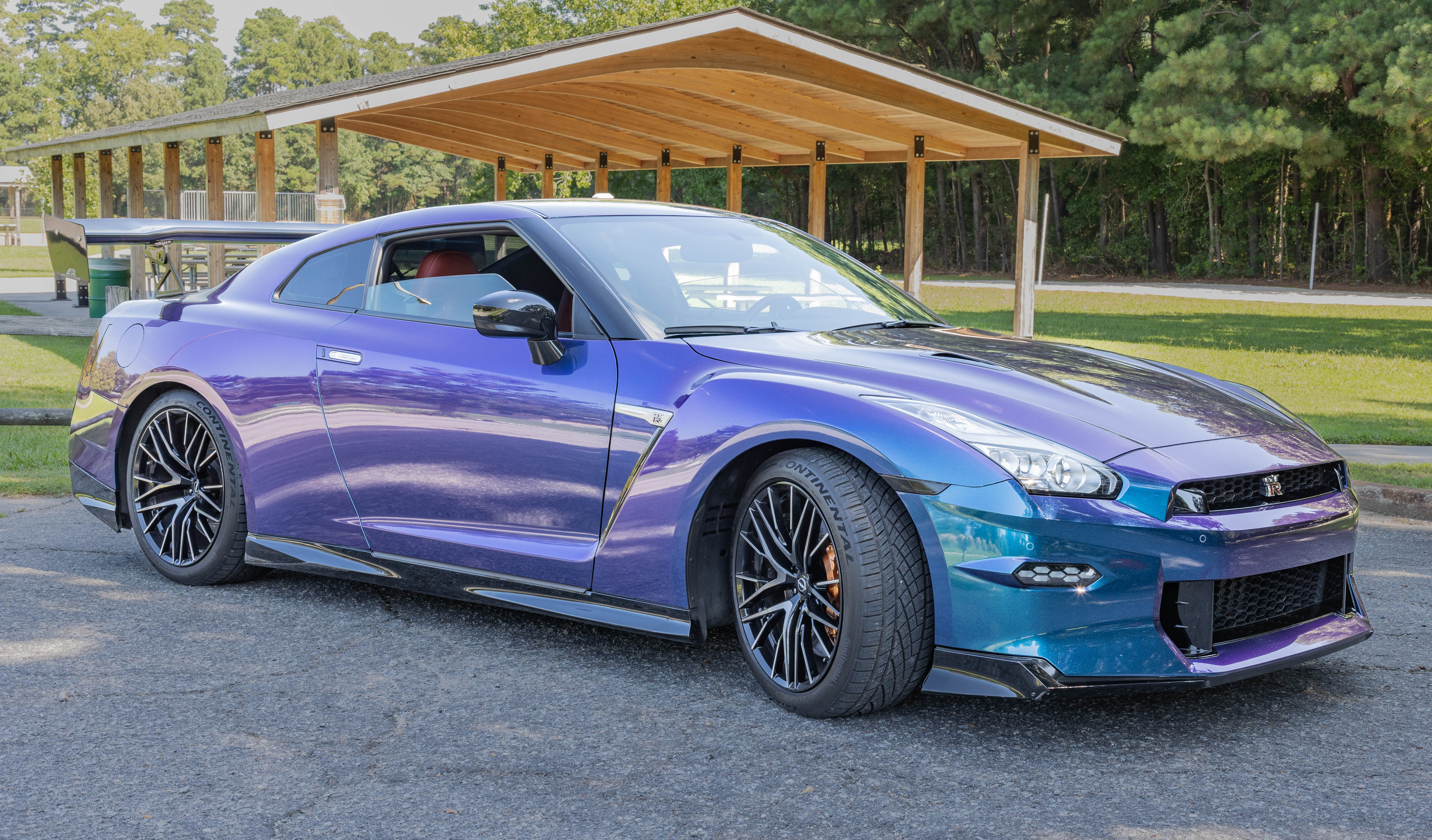 Front three-quarter view of the 2024 Nissan GT-R Premium in color-shift purple-to-blue paint parked beside a park pavilion