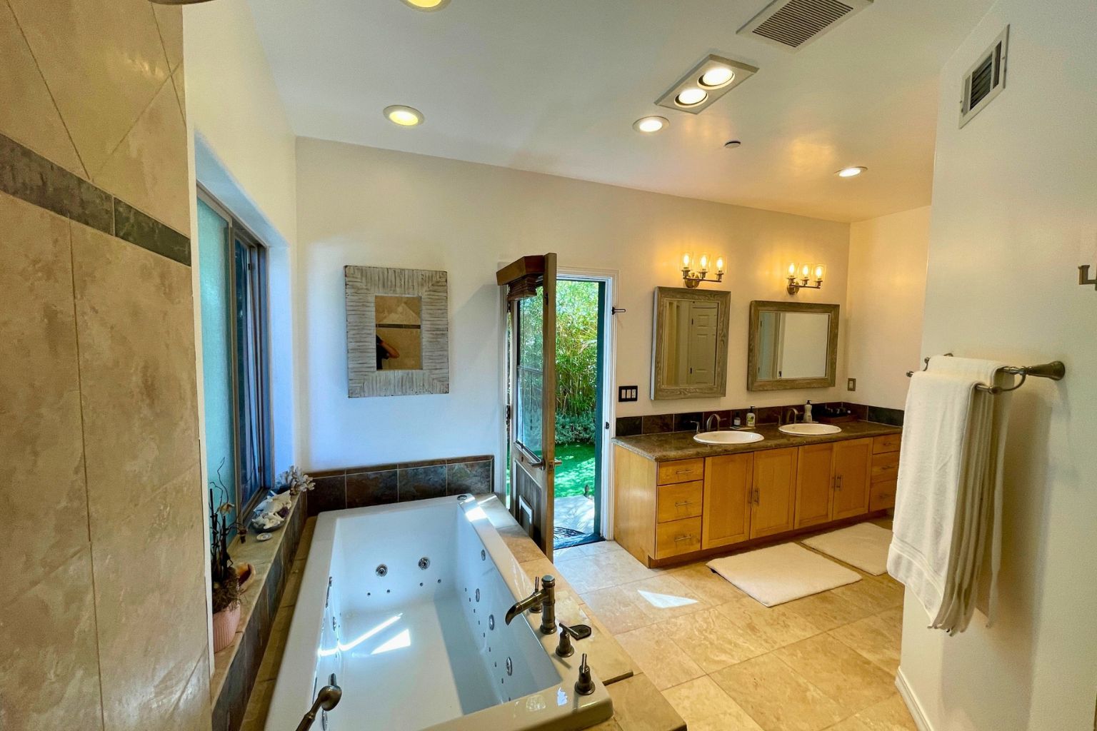 Primary bath with a travertine encased jetted tub, dual vanity, and a private door opening to the garden