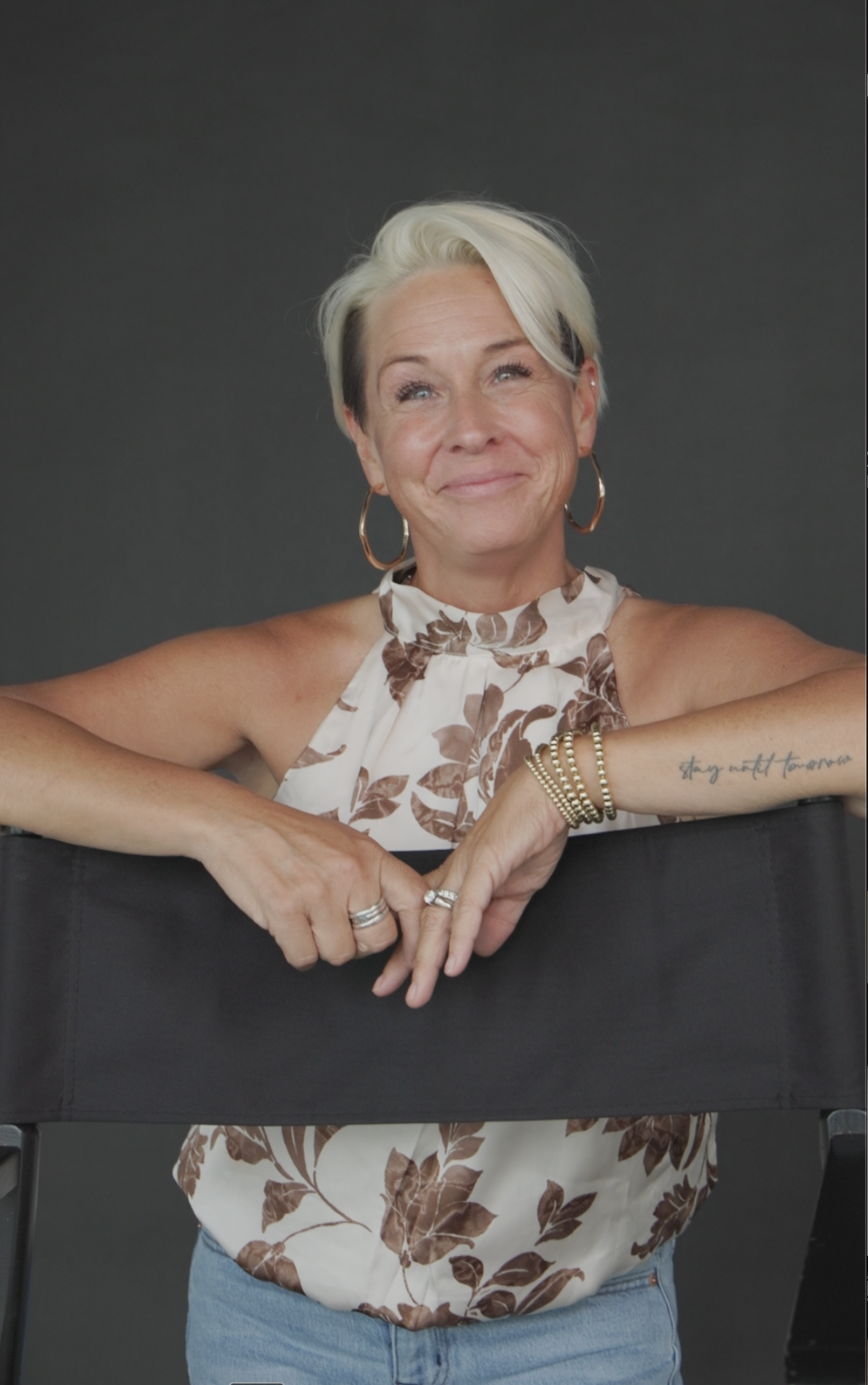 Lori smiling in a floral halter top leaning forward on a director's chair