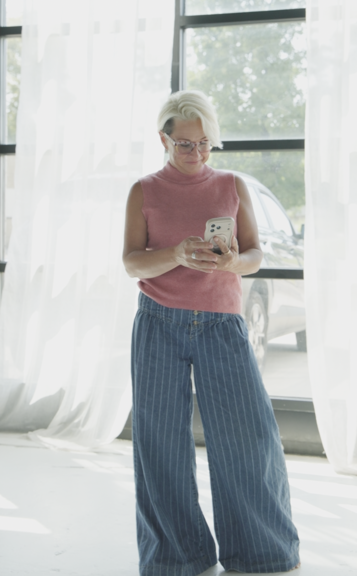 Lori looking at her phone in a pink sleeveless top and wide-leg denim pants